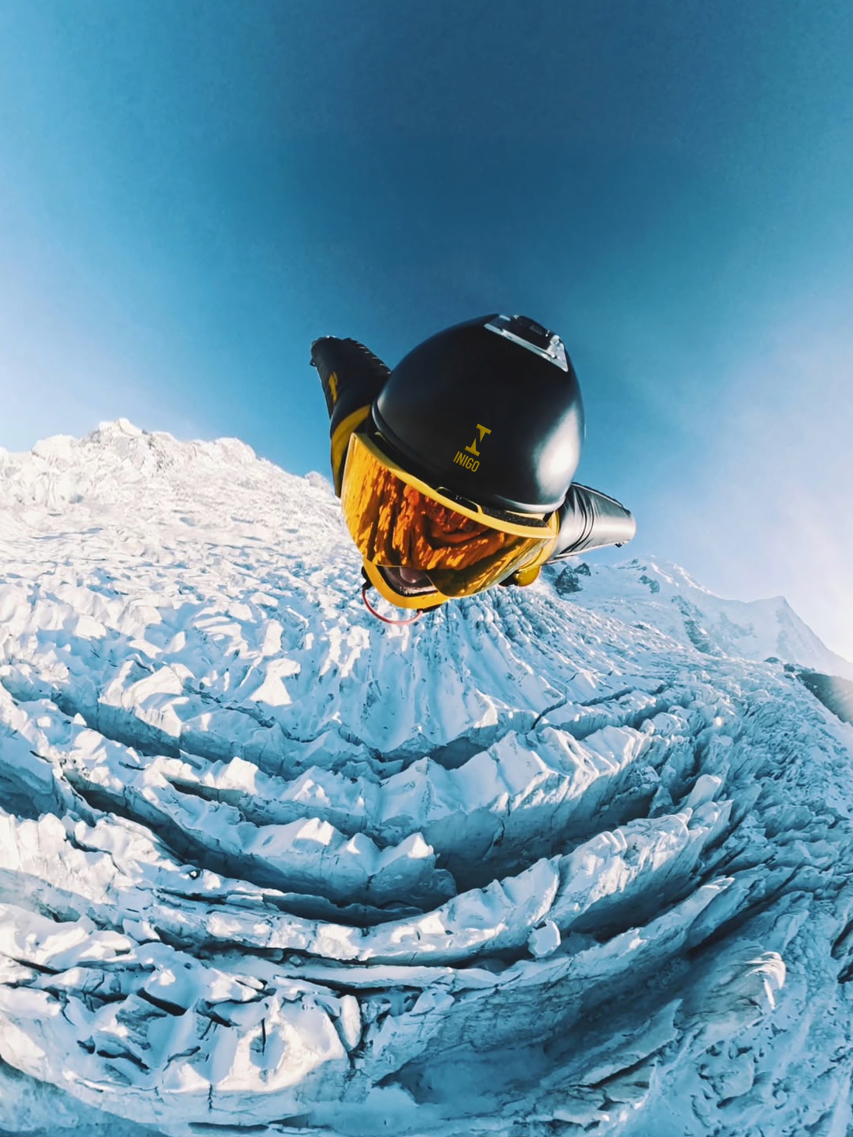 Wingsuit flyer soaring over a frozen mountain landscape, captured from above with the Inigo logo visible on the helmet.