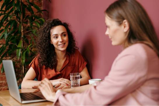 Two individuals sit at a wooden table with a laptop, having a discussion; one smiles and wears a rust-colored shirt, while the other listens attentively in a pink blouse, with a brick wall and plant in the background.