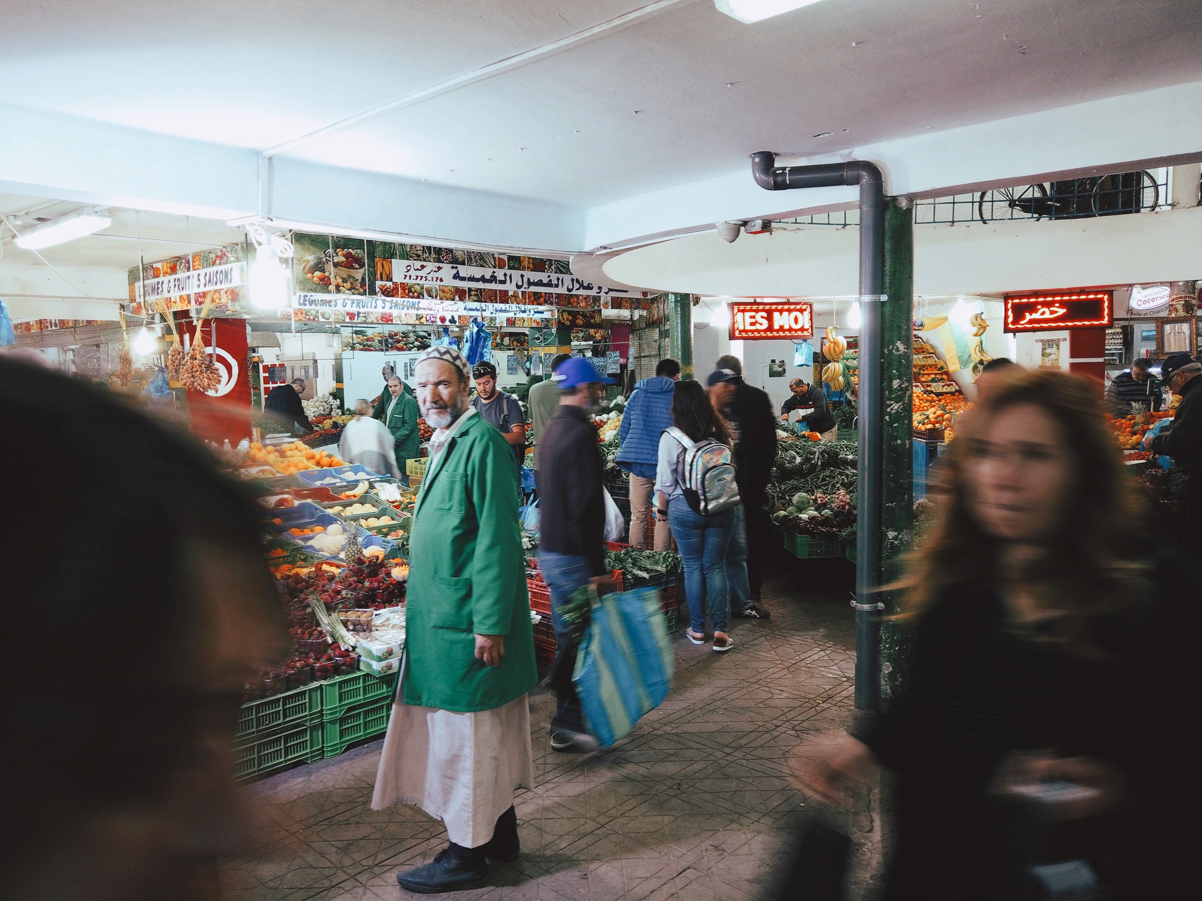 Un homme immobile avec une veste verte se tient debout dans un marché aux fruits pendant que d'autres personnes se déplacent autour