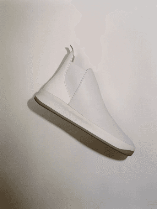 A sleek, minimalist white sneaker with a high-top design is shown against a plain background, highlighting its smooth leather finish and subtle stitching details.