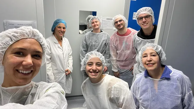 A team of researchers wearing protective lab coats and hairnets smiling inside a biotechnology facility.