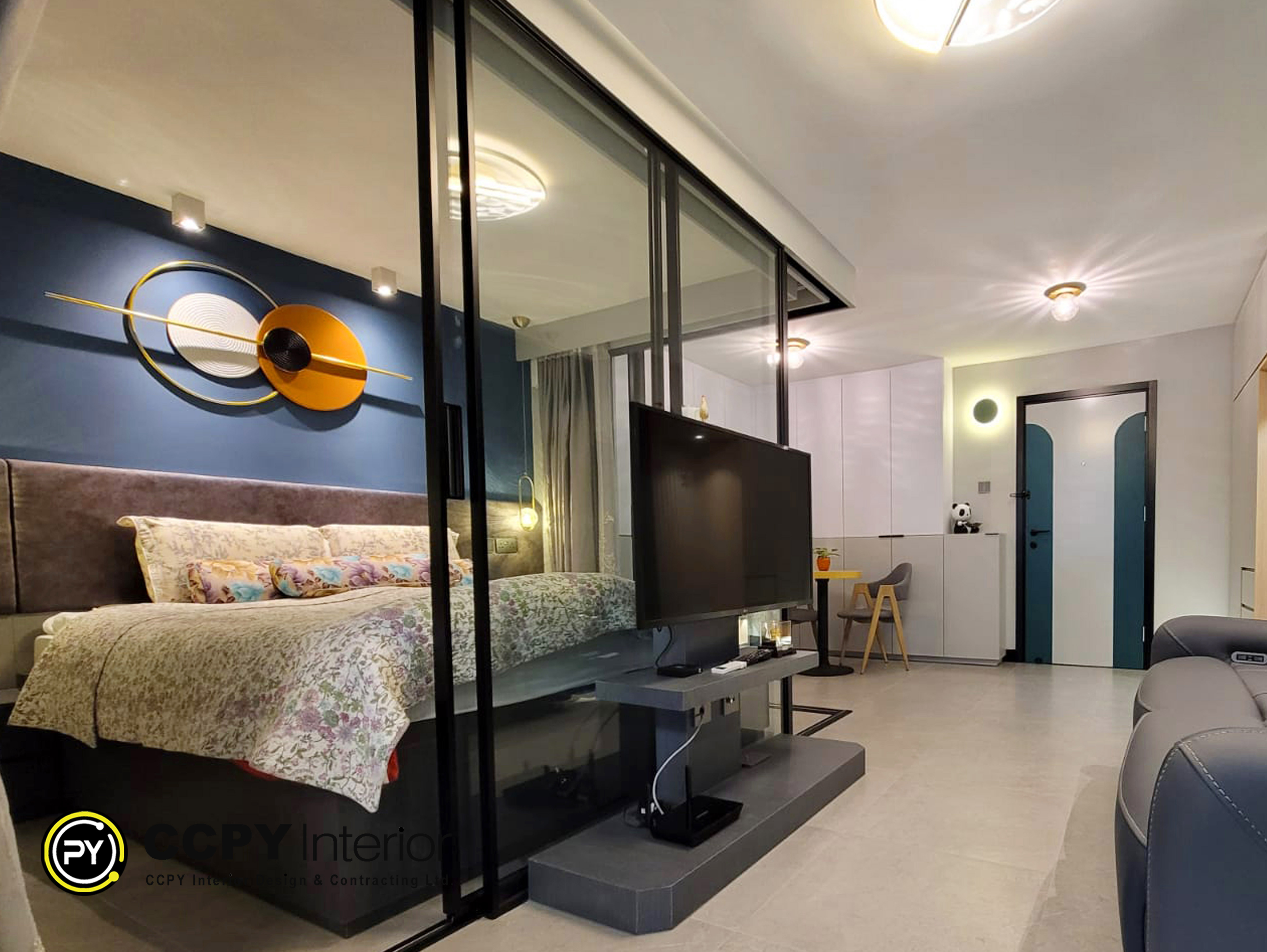 This view highlights the clever use of a glass partitioning system (強化玻璃隔斷牆) with black frames to create a sleek divide between the bedroom and living area. The TV console is mounted directly to the partition structure, an efficient use of space.