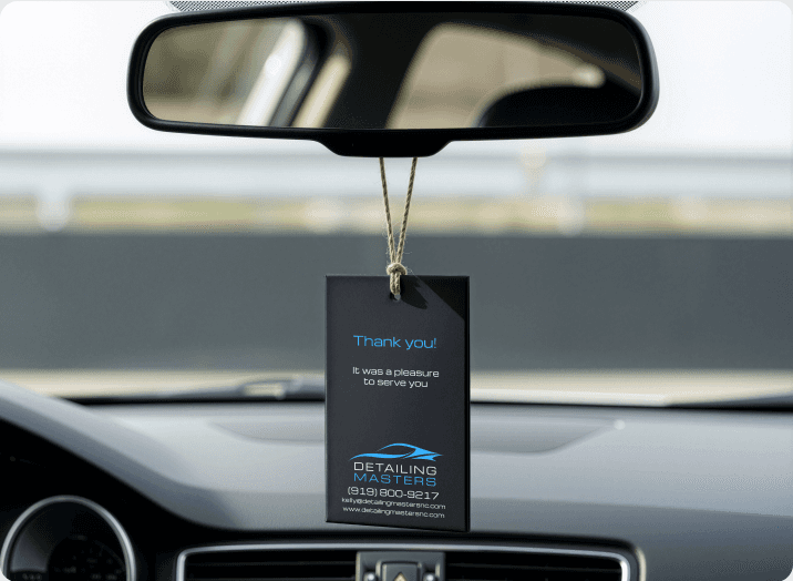 Photo of a hanging tag showing the Detailing Masters logo and "thank you" hanging from the rear view mirror on the interior of an automobile