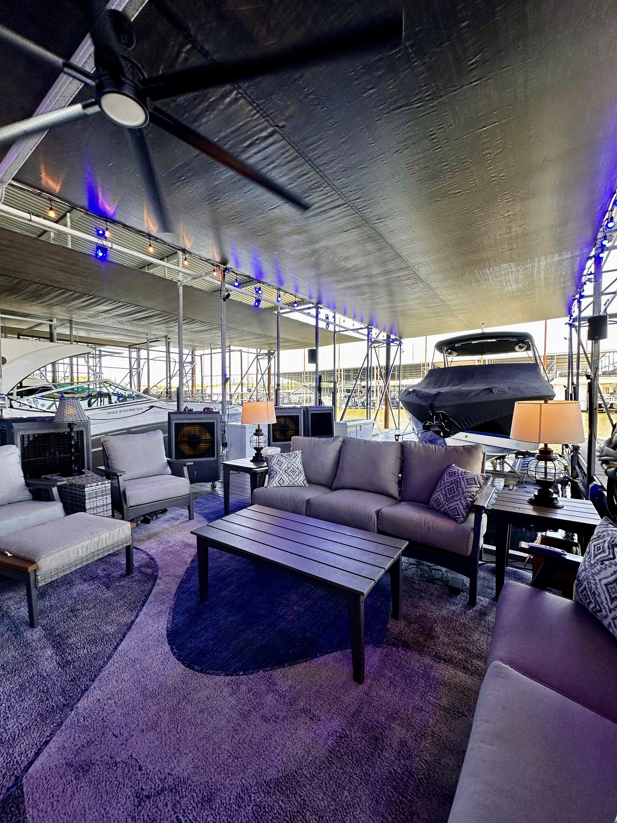 A covered and furnished outdoor seating area on a boat dock featuring several cushioned chairs, a coffee table, a large ceiling fan, and overhead lighting, with a luxury boat docked beside it.