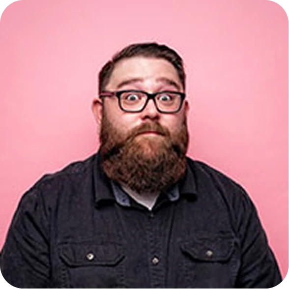 Studio headshot portrait of a man with glasses and a full beard posing against a solid pink backdrop.
