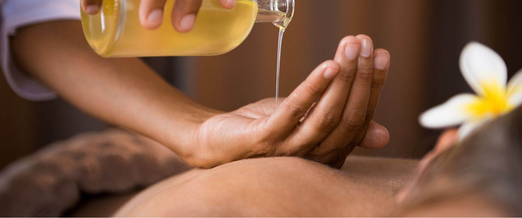 A person's hand pours warm oil from a clear bottle onto another person's back, illustrating the practice of Abhyanga massage, known for its relaxation and health benefits.