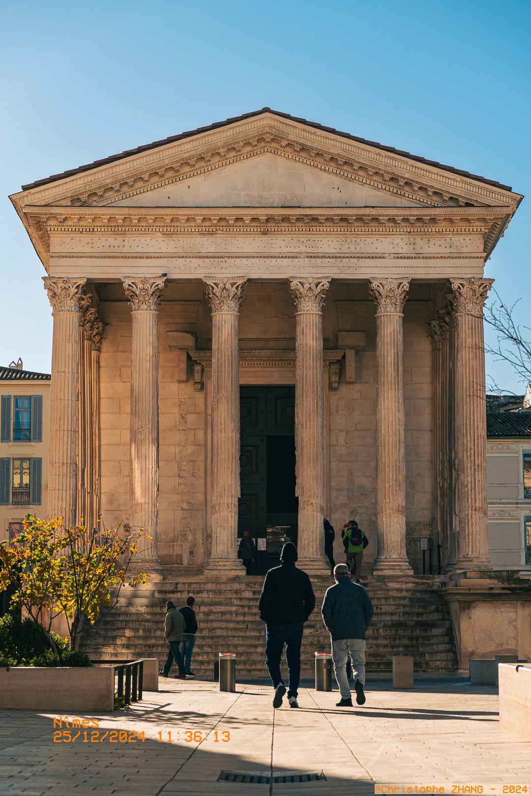 Entrée de la Maison Carrée à Nîmes, avec des visiteurs montant les marches sous un ciel bleu.