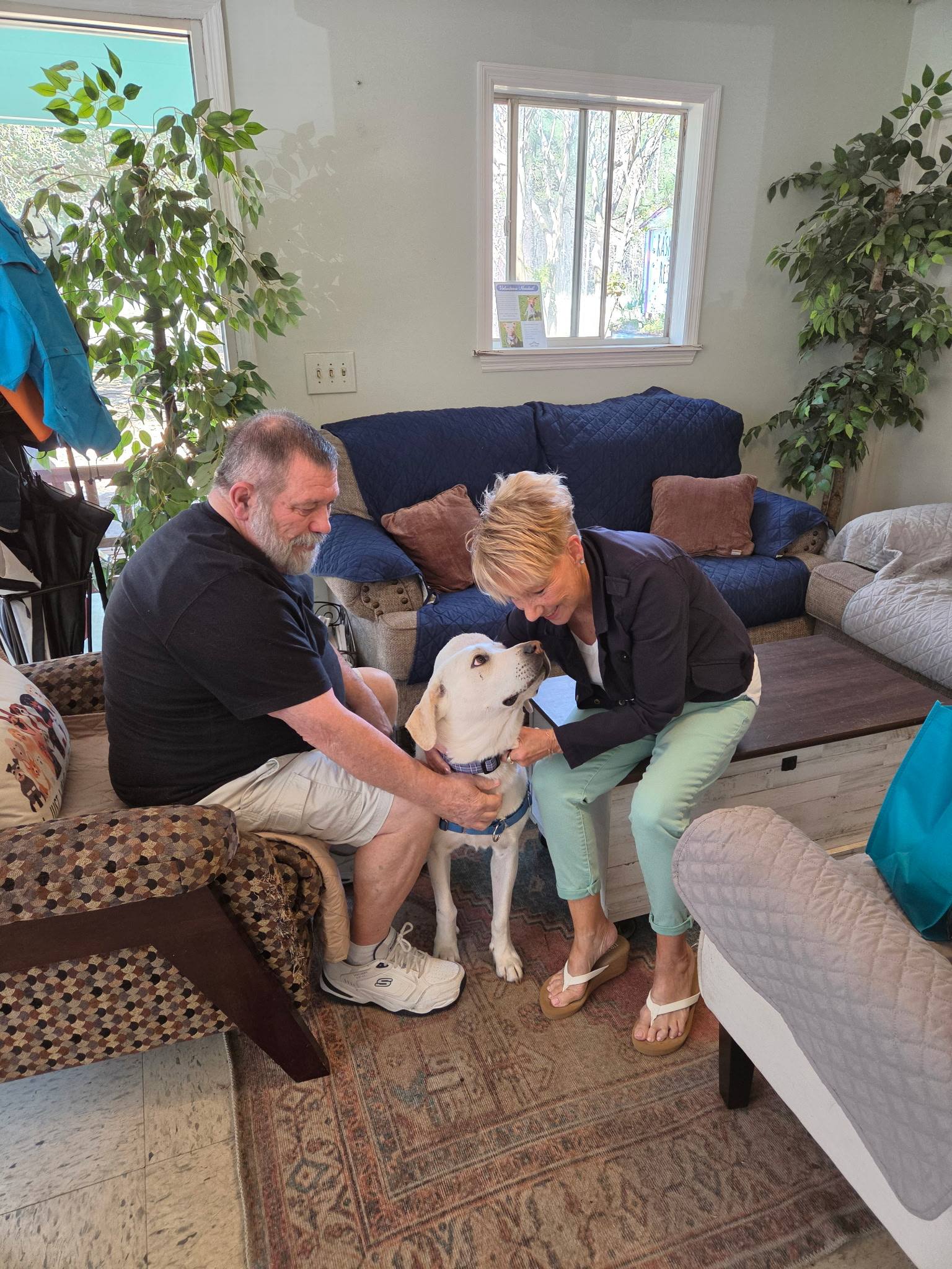 A man and a woman sit on a couch, interacting with a small dog on a table in a cozy living room setting.