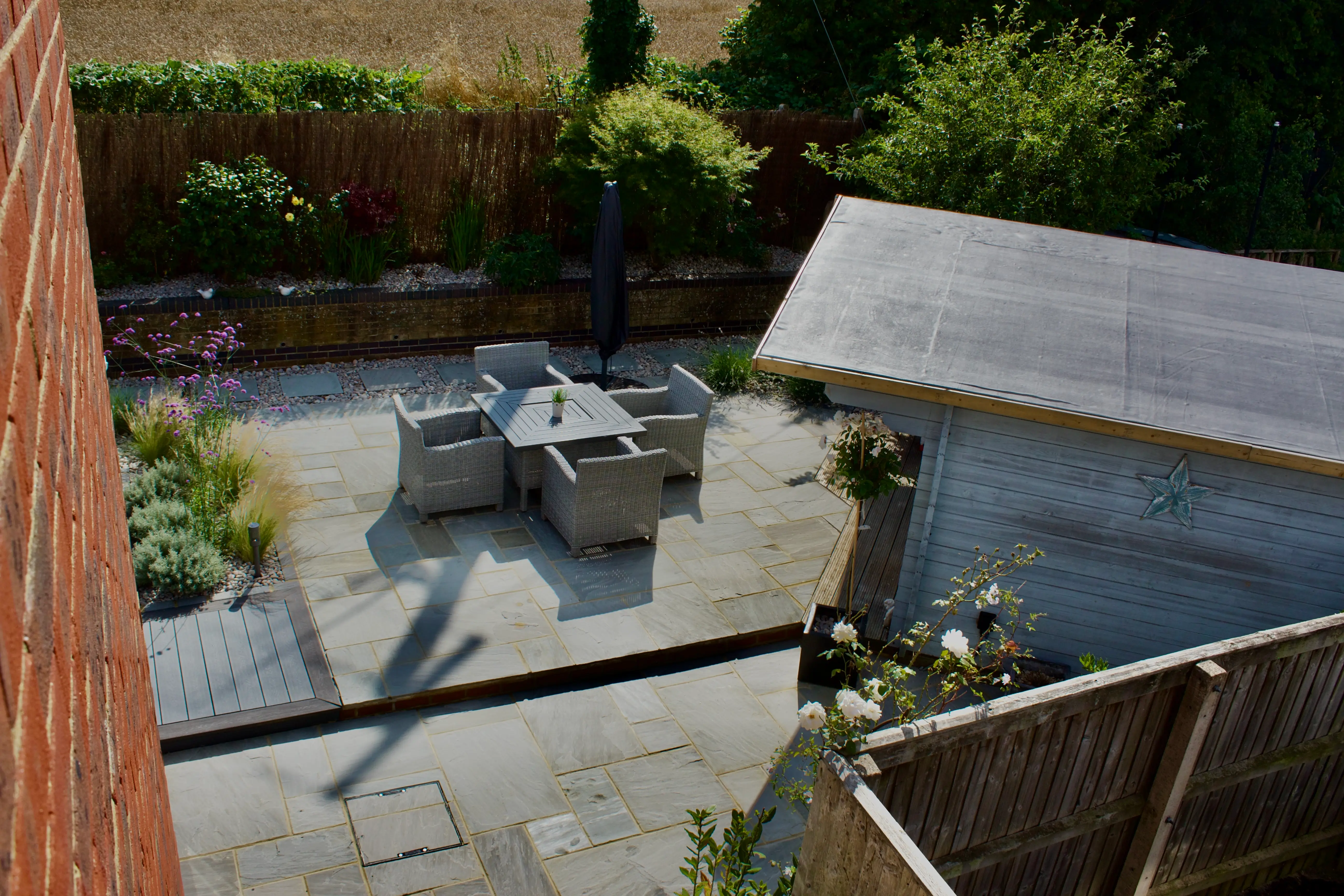 A view of a patio with a table and chairs, surrounded by greenery and a fence, under bright sunlight.