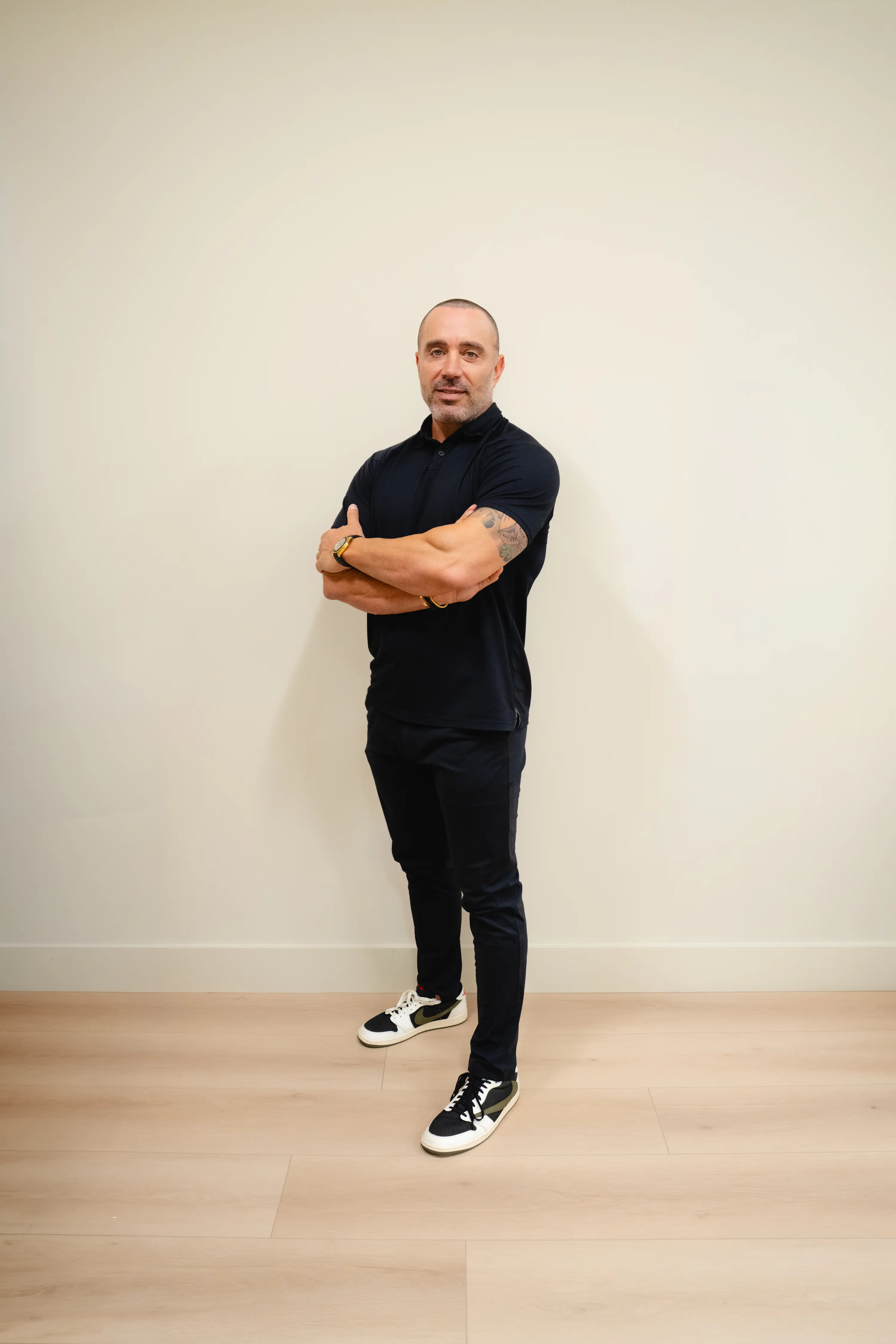 Man in a black polo standing with arms crossed against a white wall