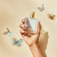 Hand holding a glass perfume bottle surrounded by blue butterflies against beige background