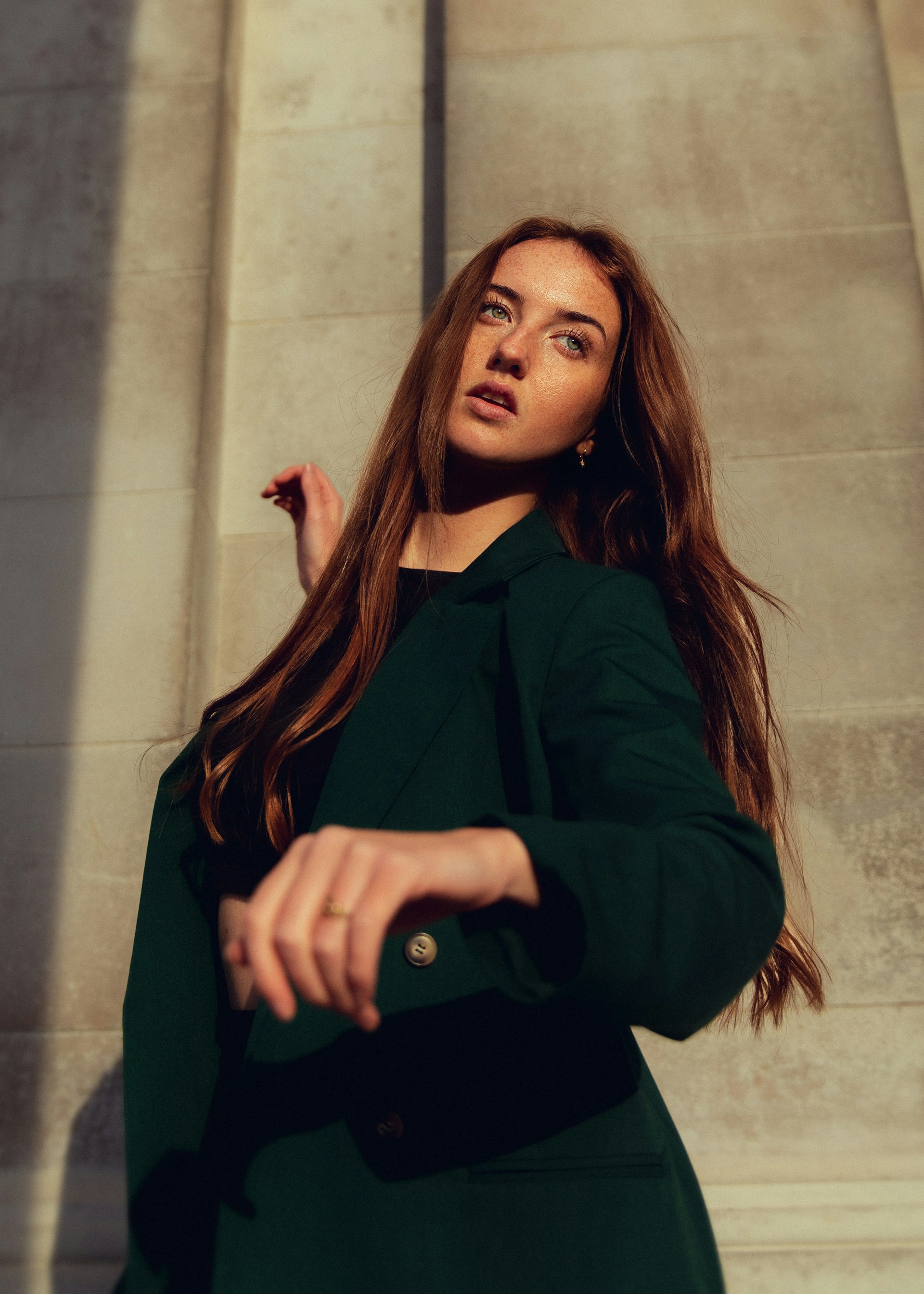 A profile shot of the person leaning against a white brick wall, looking off to the side. They are wearing a dark grey jacket and a black beanie, against a background of green foliage.