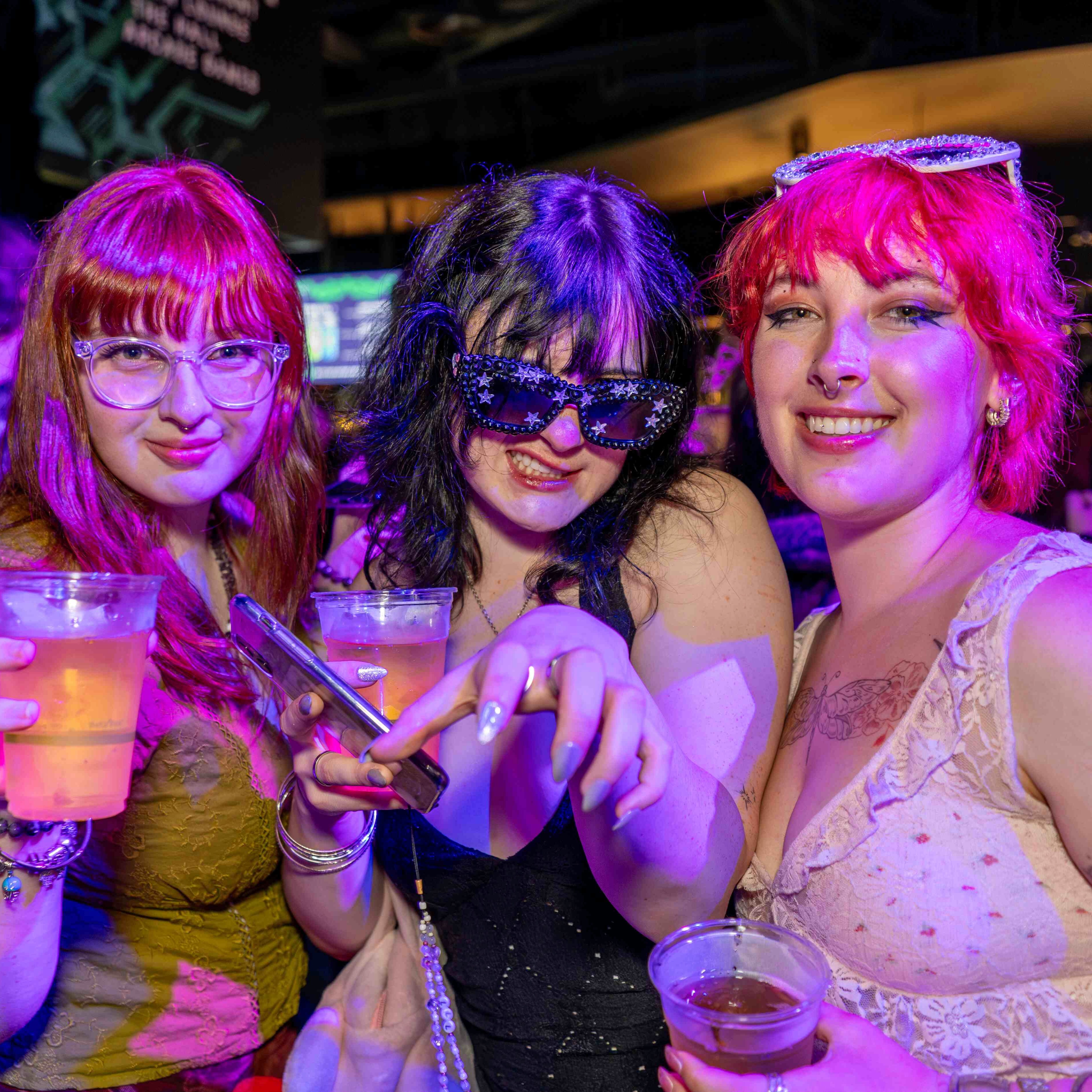 three girls under pink lights toasting the camera and smiling