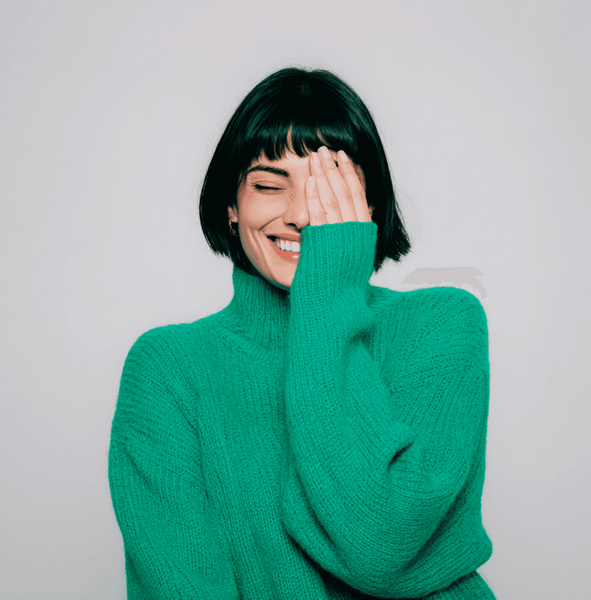 A woman with short black hair and green sweater smiles, covering her face with her hand.