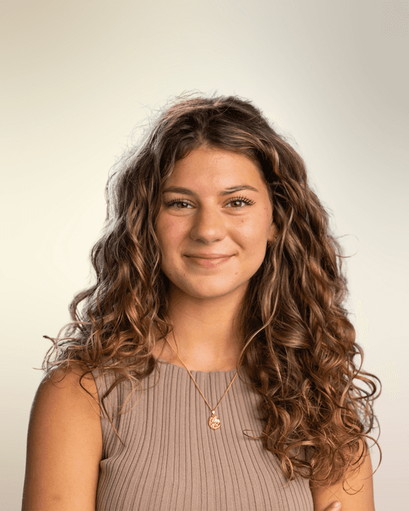Portrait of Alexis Hedley, Campaign Manager at Playkit, smiling in front of a beige wall and wearing a black strapless top with a gold cross necklace.