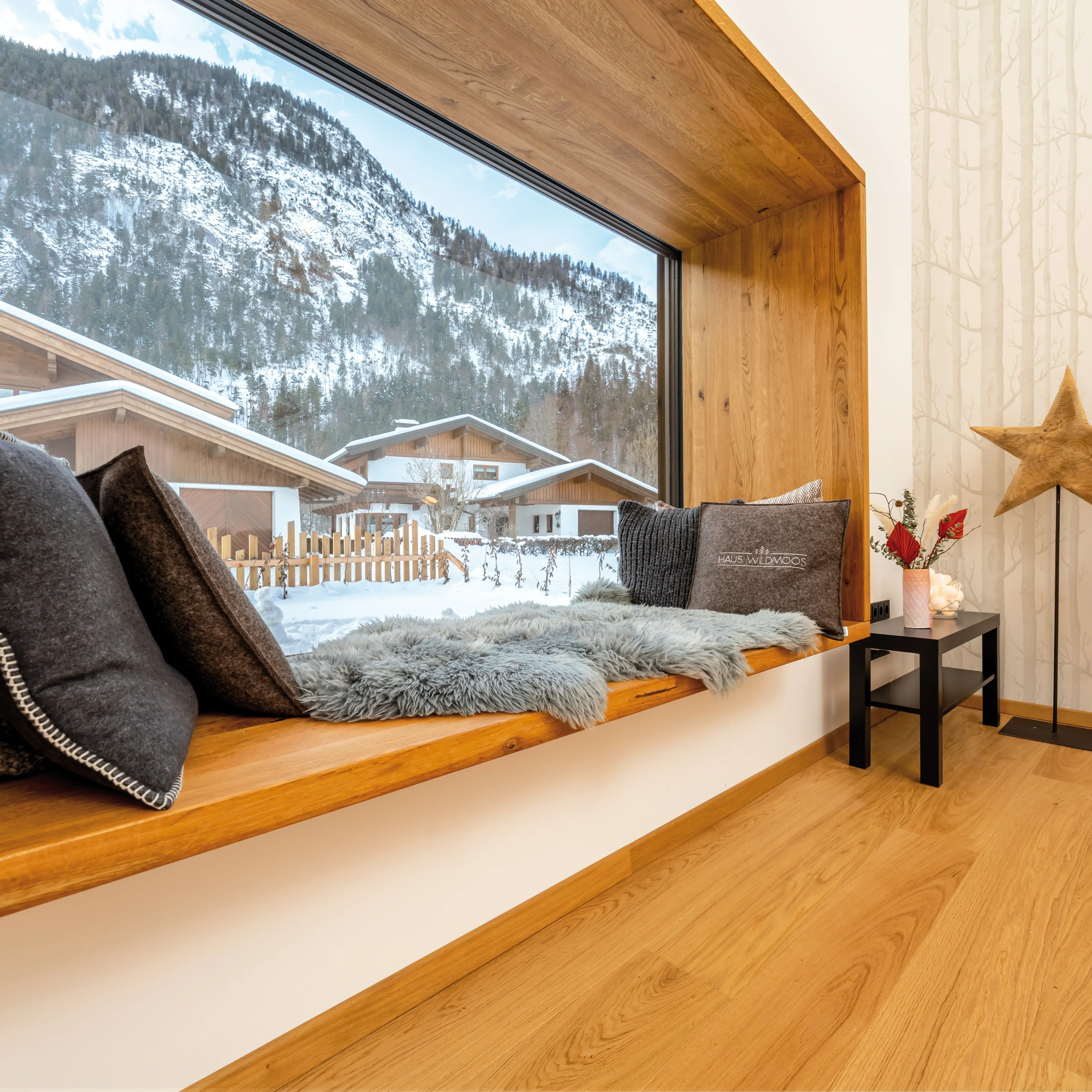Gemütliche Fensterbank mit Kissen vor einem verschneiten Bergblick in stilvollem Alpenchalet-Ambiente.