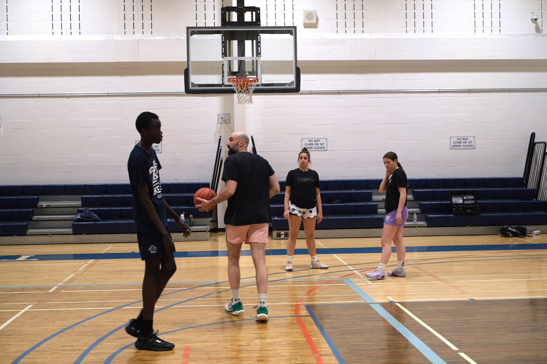 A youth basketball player in an orange jersey dribbling aggressively past a defender during a game, with a referee and spectators visible in the background on an indoor court.
