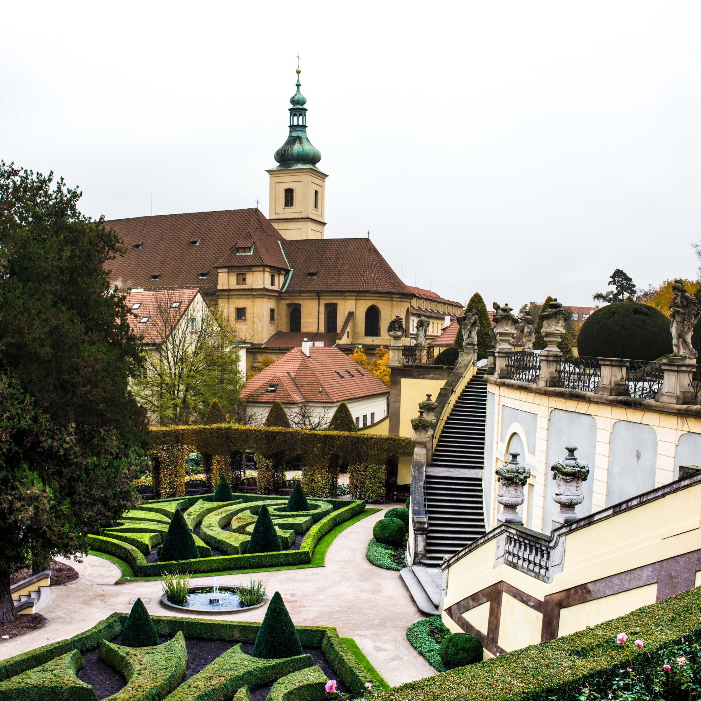 A large yellow palace with a green-domed tower overlooks a formal garden with manicured hedges and stone stairs.