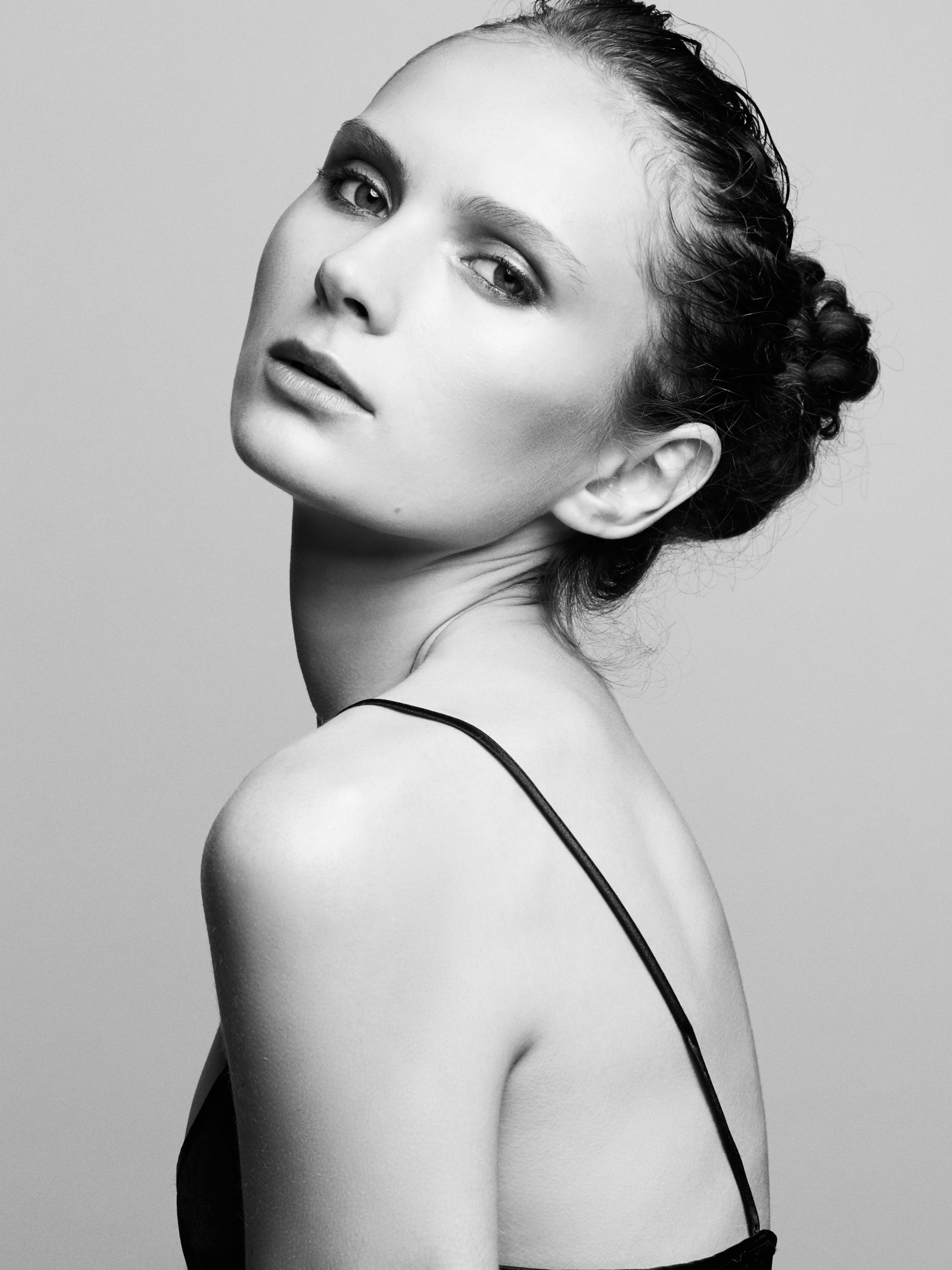 Upper body black and white portrait of a female model sitting with her dark hair tied into a bun and her body turned away with her face turned back to camera and wearing a dark string strapped silk singlet.