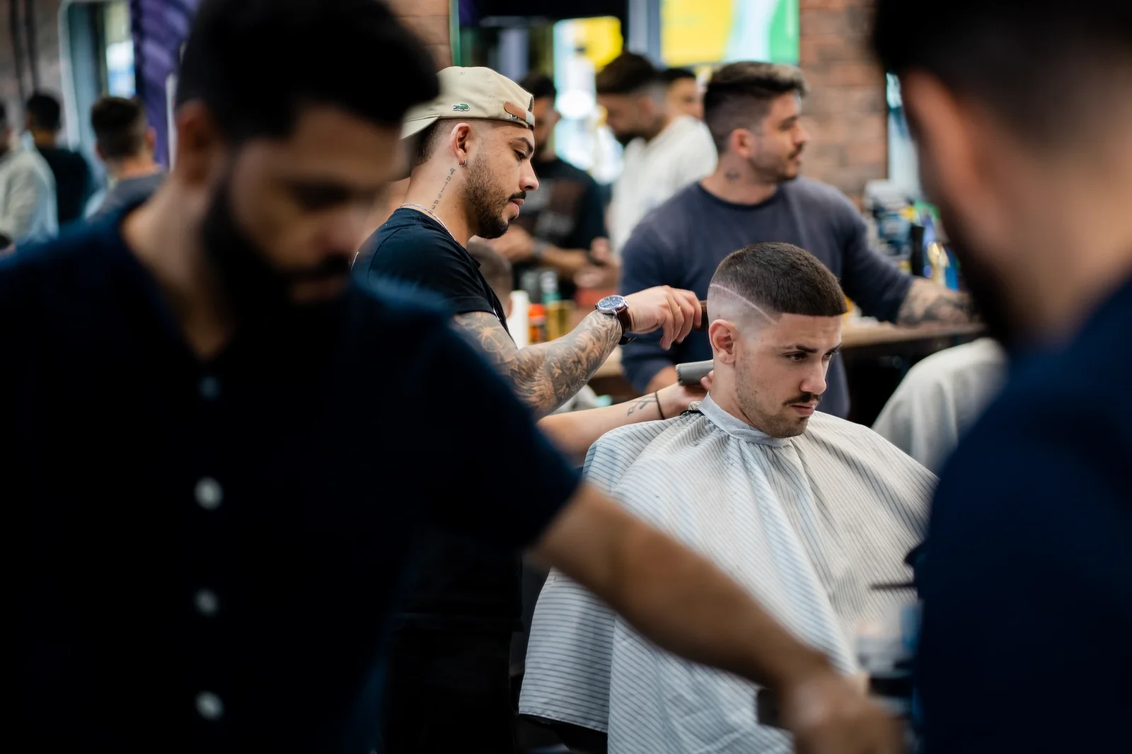 Barber giving a fade haircut to a client in a busy barbershop with multiple customers and staff present.