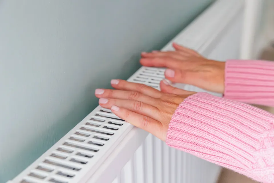 A homeowner in a pink jumper pressing hands against a lukewarm panel radiator connected to a heat pump system in a UK home.