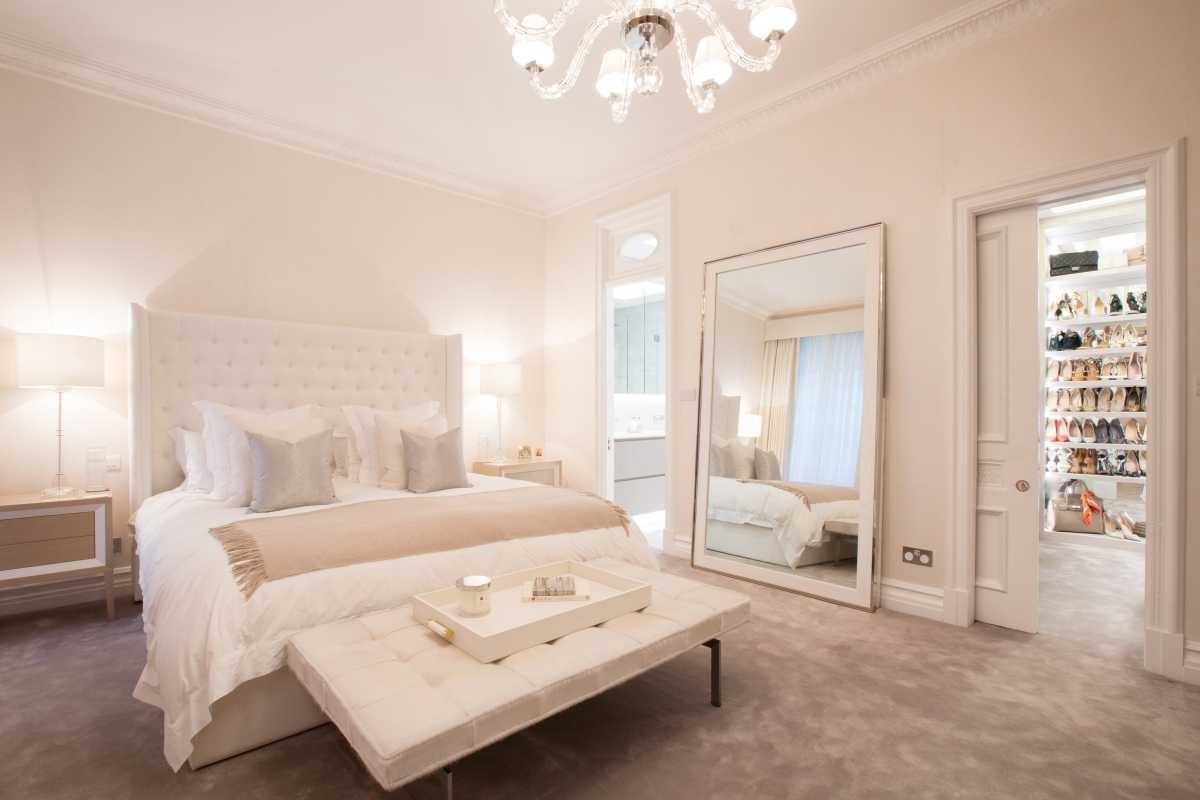 Elegant master bedroom interior designed by Doll & Co. showing a tufted bed, full-length floor mirror, and walk-in closet entryway.