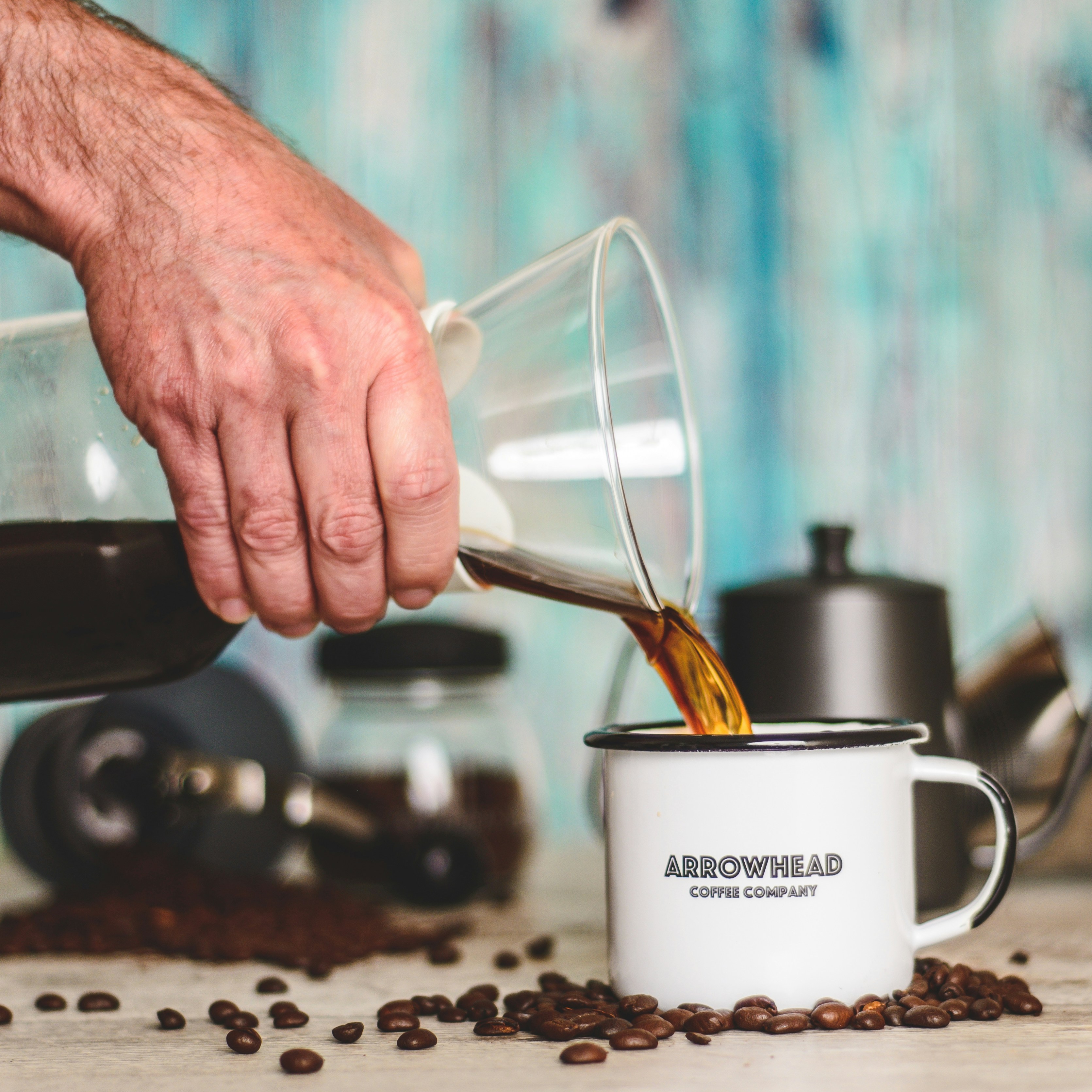 A hand pouring freshly brewed coffee into a white Arrowhead Coffee Company mug surrounded by coffee beans, symbolising creativity and focus.