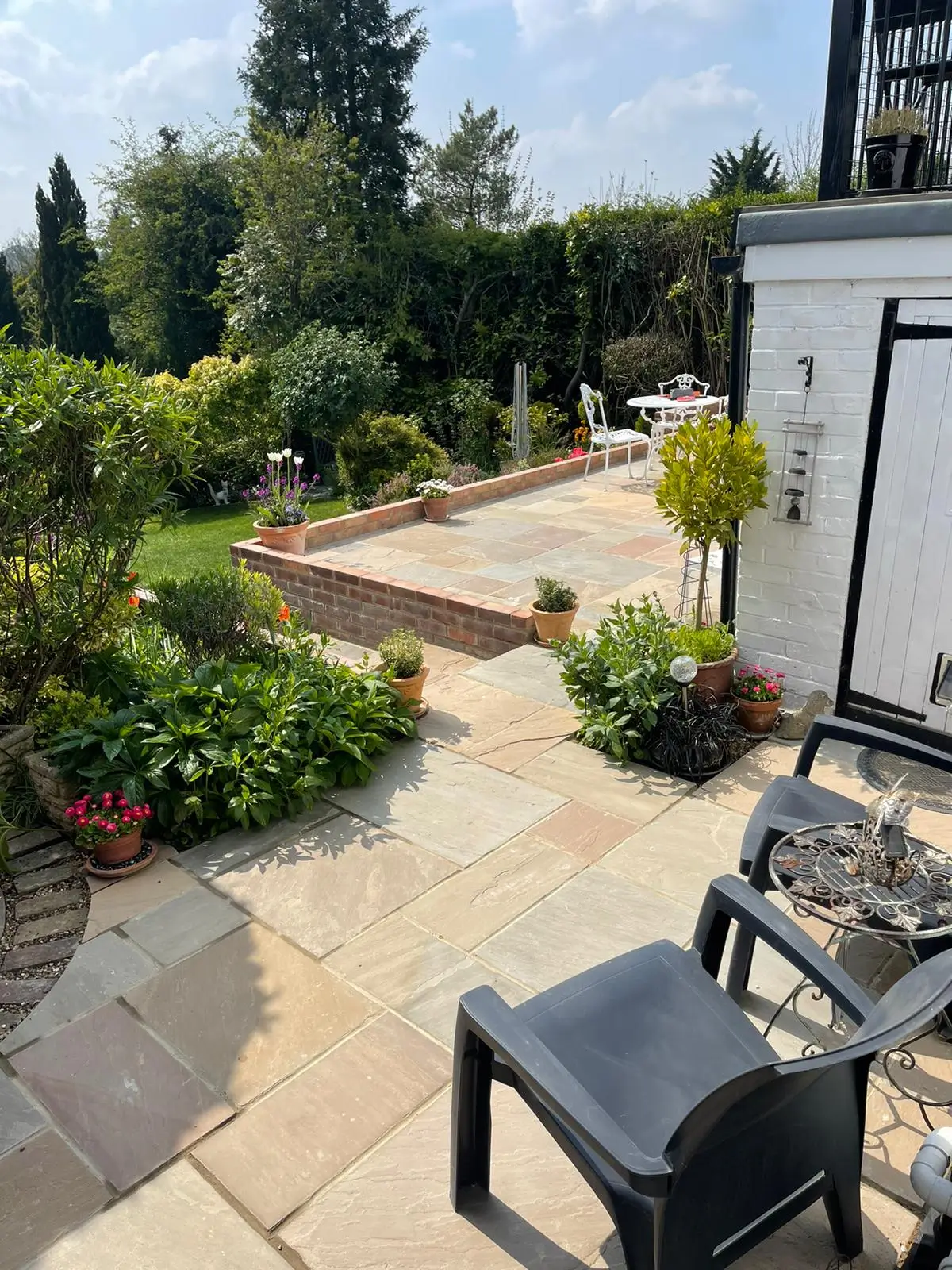 A sunny garden path with greenery, leading towards a house, featuring a black chair in the foreground.