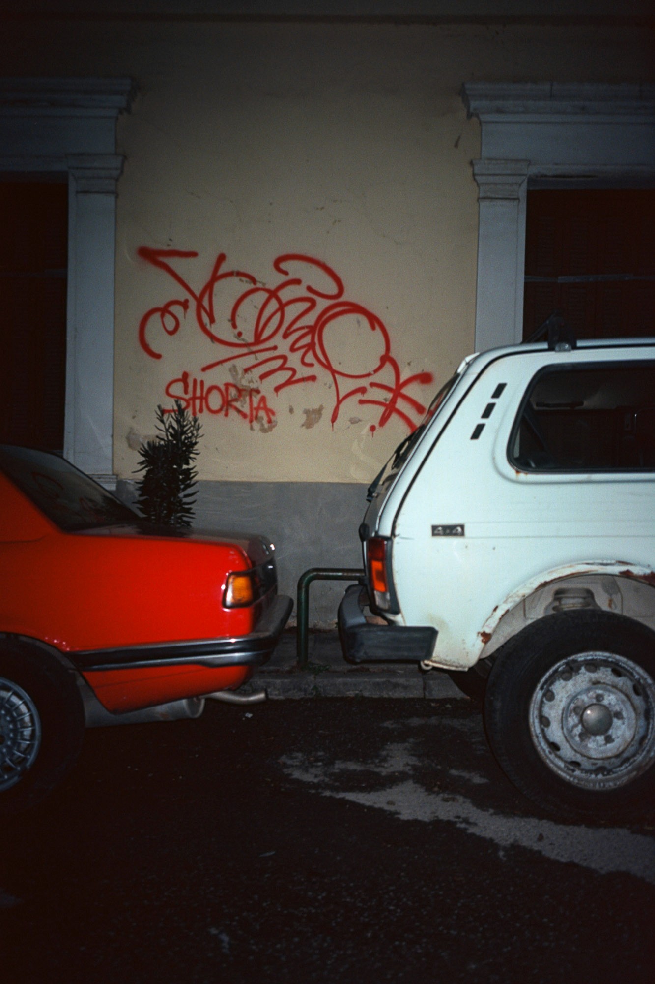 A red car and a white SUV are parked closely in front of a wall with bold red graffiti, in an urban street setting.
