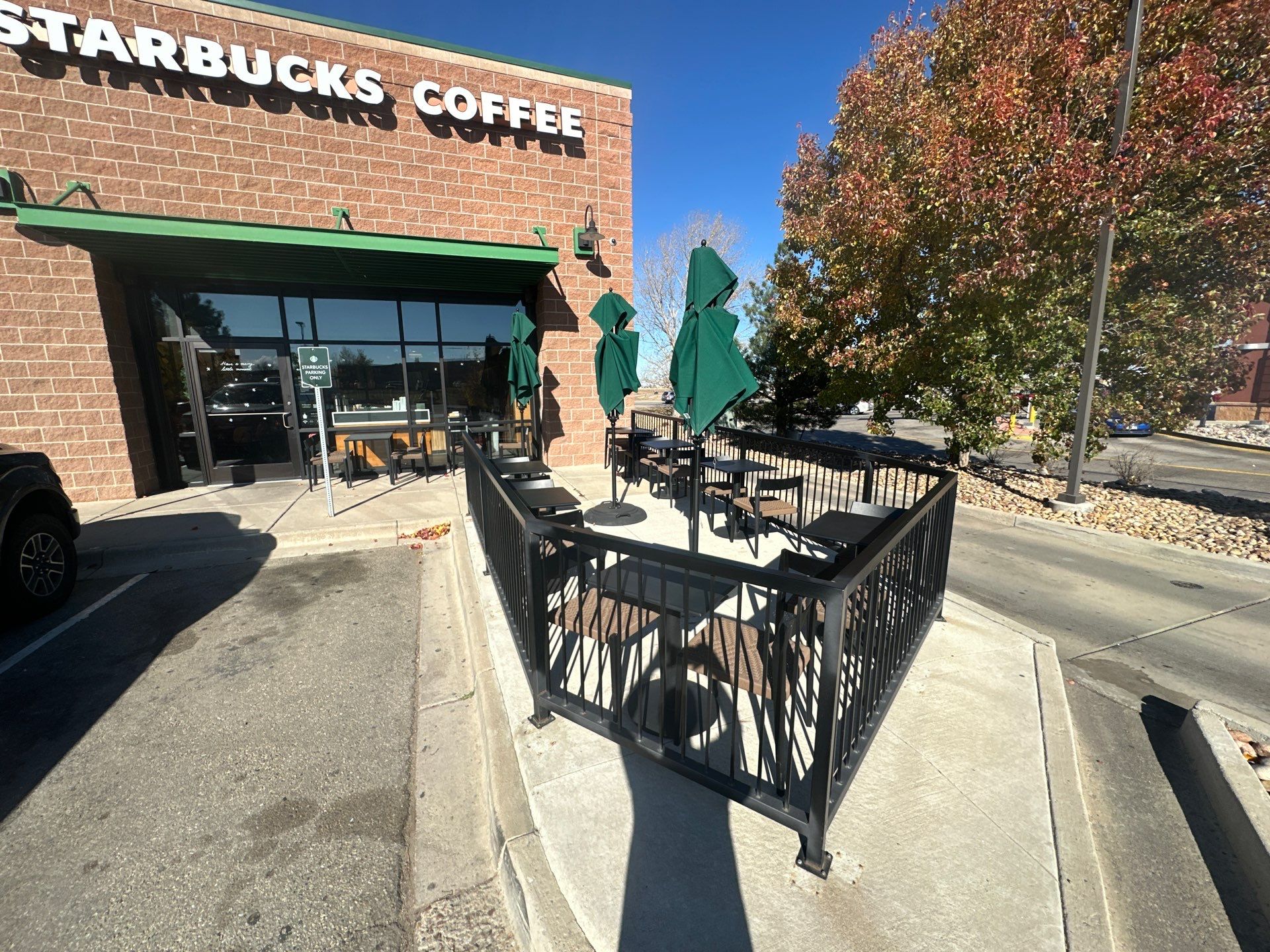 Outdoor seating area at a Starbucks location with black metal railing, umbrellas, and neatly arranged tables under clear blue skies.