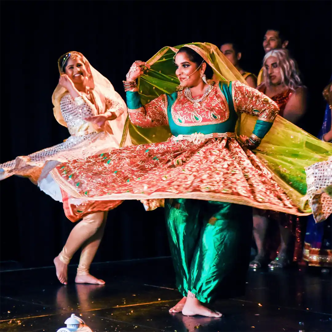 Two women in vibrant traditional South Asian attire dance on stage, spinning gracefully with flowing skirts and dupattas, whilst others in colourful costumes watch in the background.