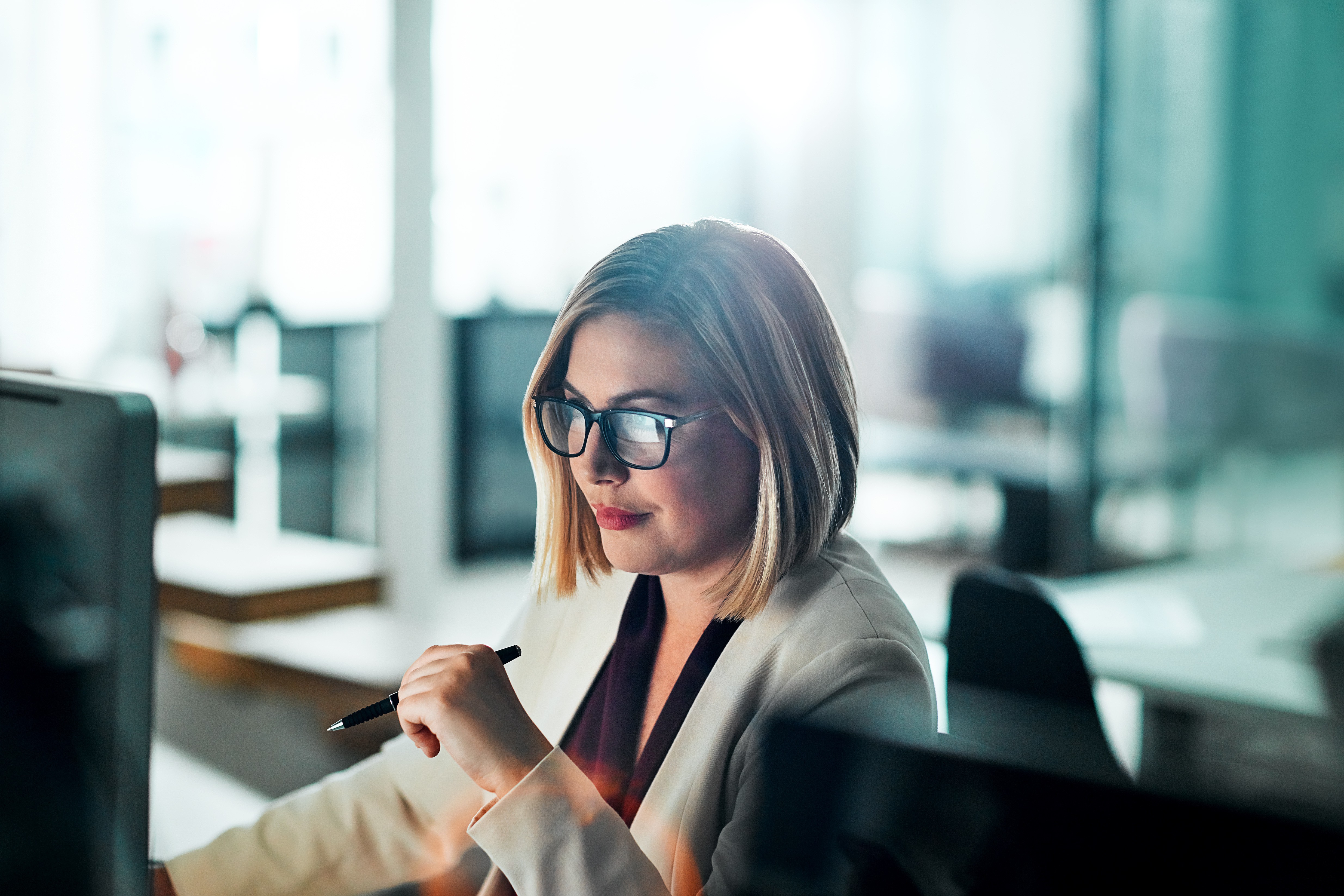 Geschäftsfrau mit Brille arbeitet konzentriert am Computer in einem modernen Büro und hält einen Stift in der Hand.