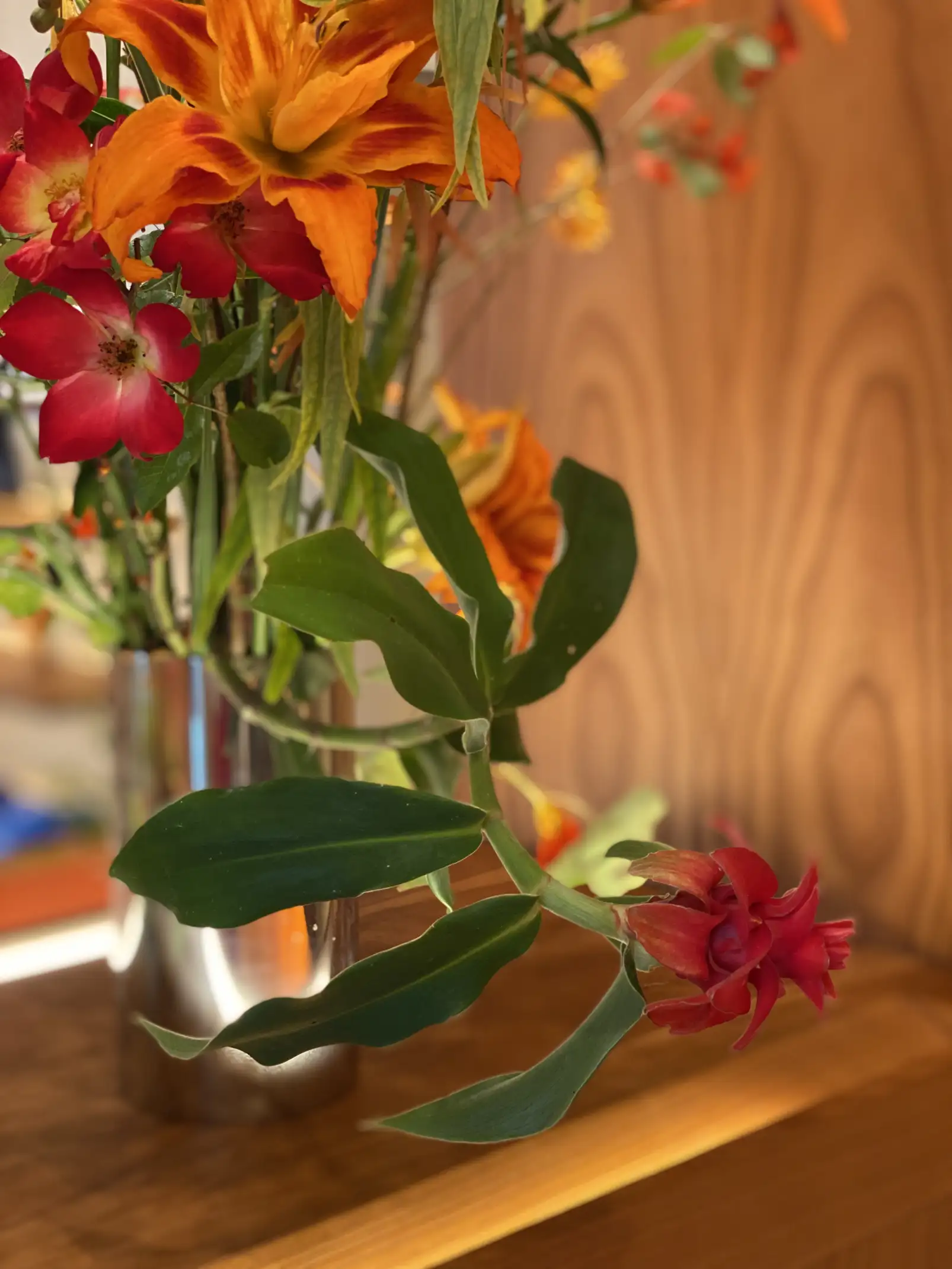 Detalle de un arreglo floral en jarrón metálico con lirios naranjas, flores rojas y hojas verdes brillantes que sobresalen del conjunto. El fondo de madera cálida resalta la intensidad cromática de las flores, creando una composición elegante y vibrante.