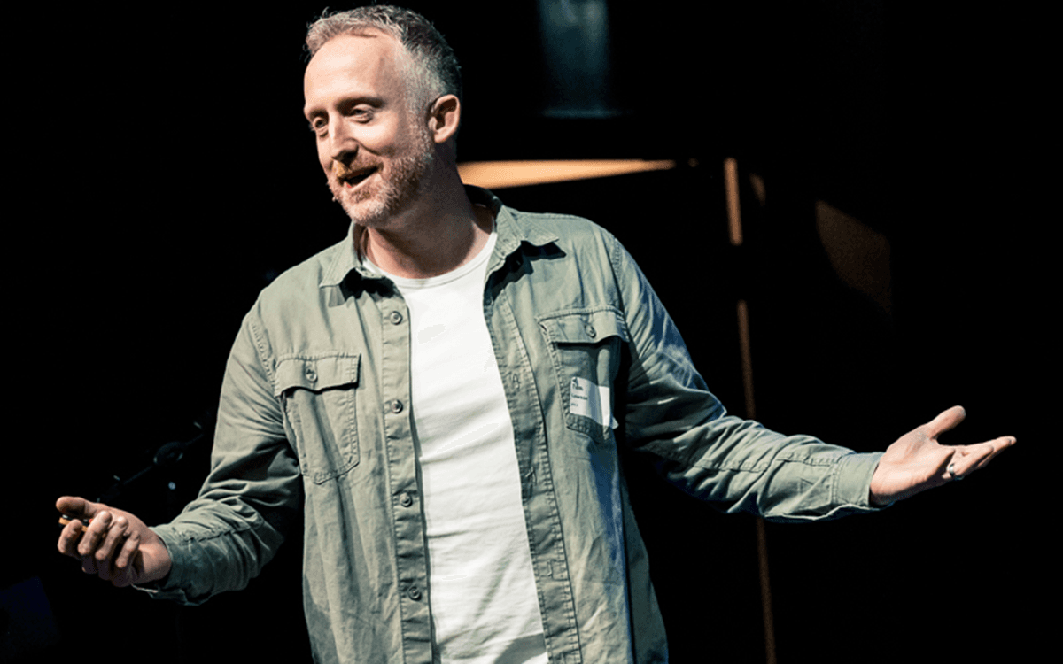 Person standing on stage with arms slightly outstretched, wearing a light green button-up shirt over a white t-shirt, holding a small object in one hand.