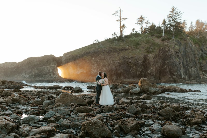 Couple in wedding attire on a rocky shoreline with golden light through a sea arch
