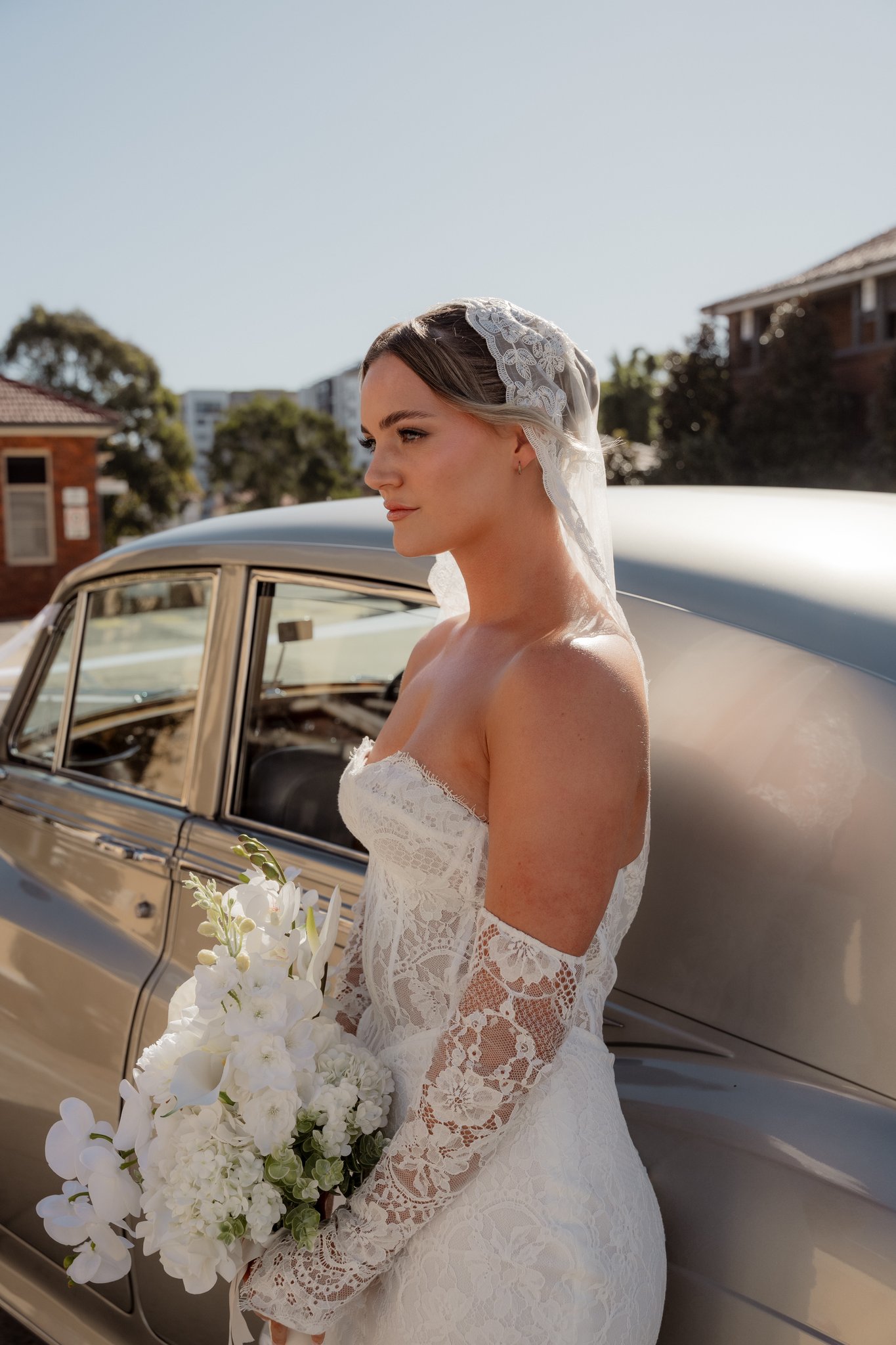 Bride in lace gown and veil holds white bouquet beside a vintage car in bright daytime sun.