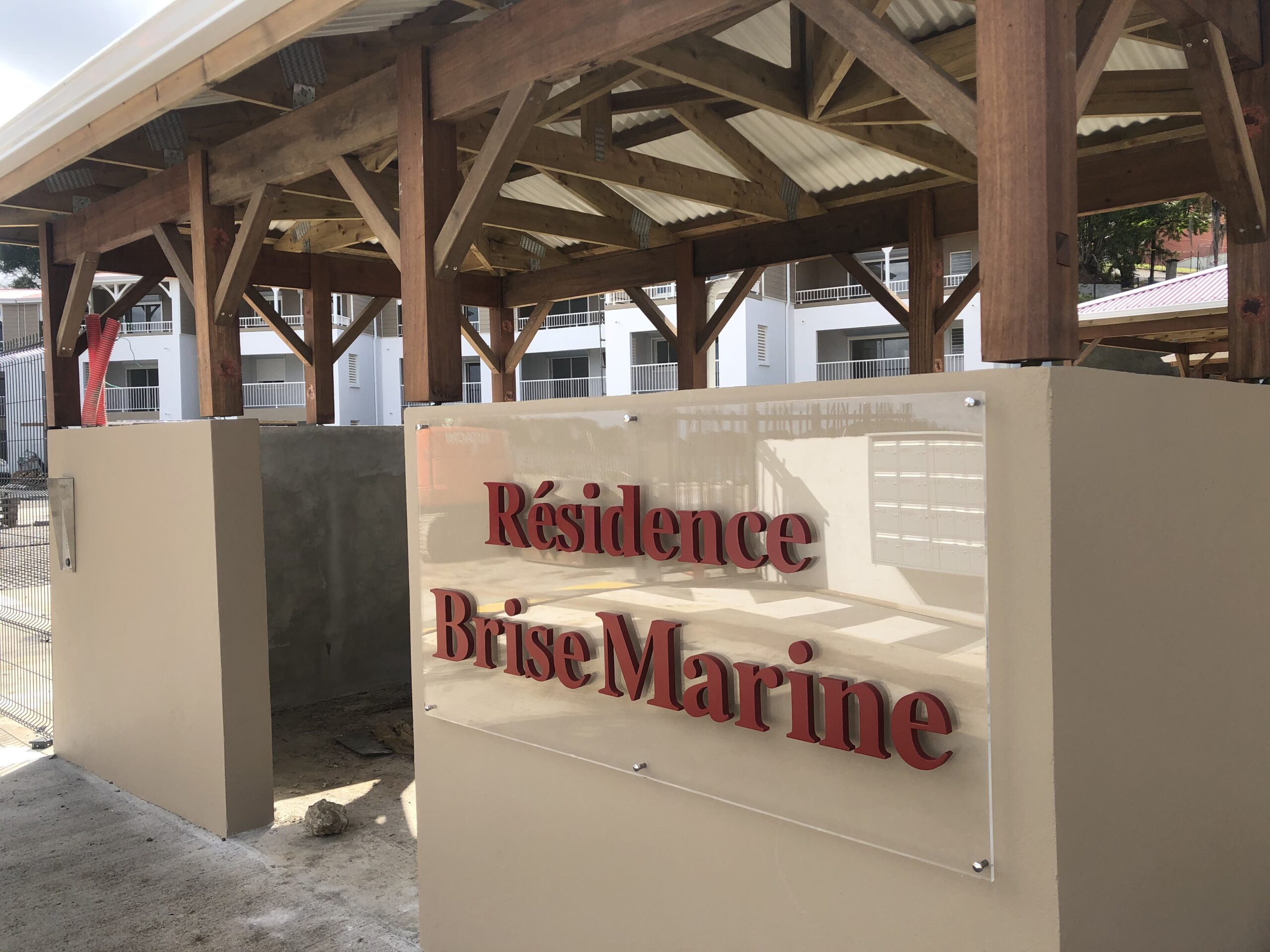 Vue extérieure de l'entrée d'un immeuble portant l'inscription « Résidence Brise Marine » en lettres rouges grasses sur un panneau beige. Des poutres en bois et des appartements modernes sont visibles à l'arrière-plan.