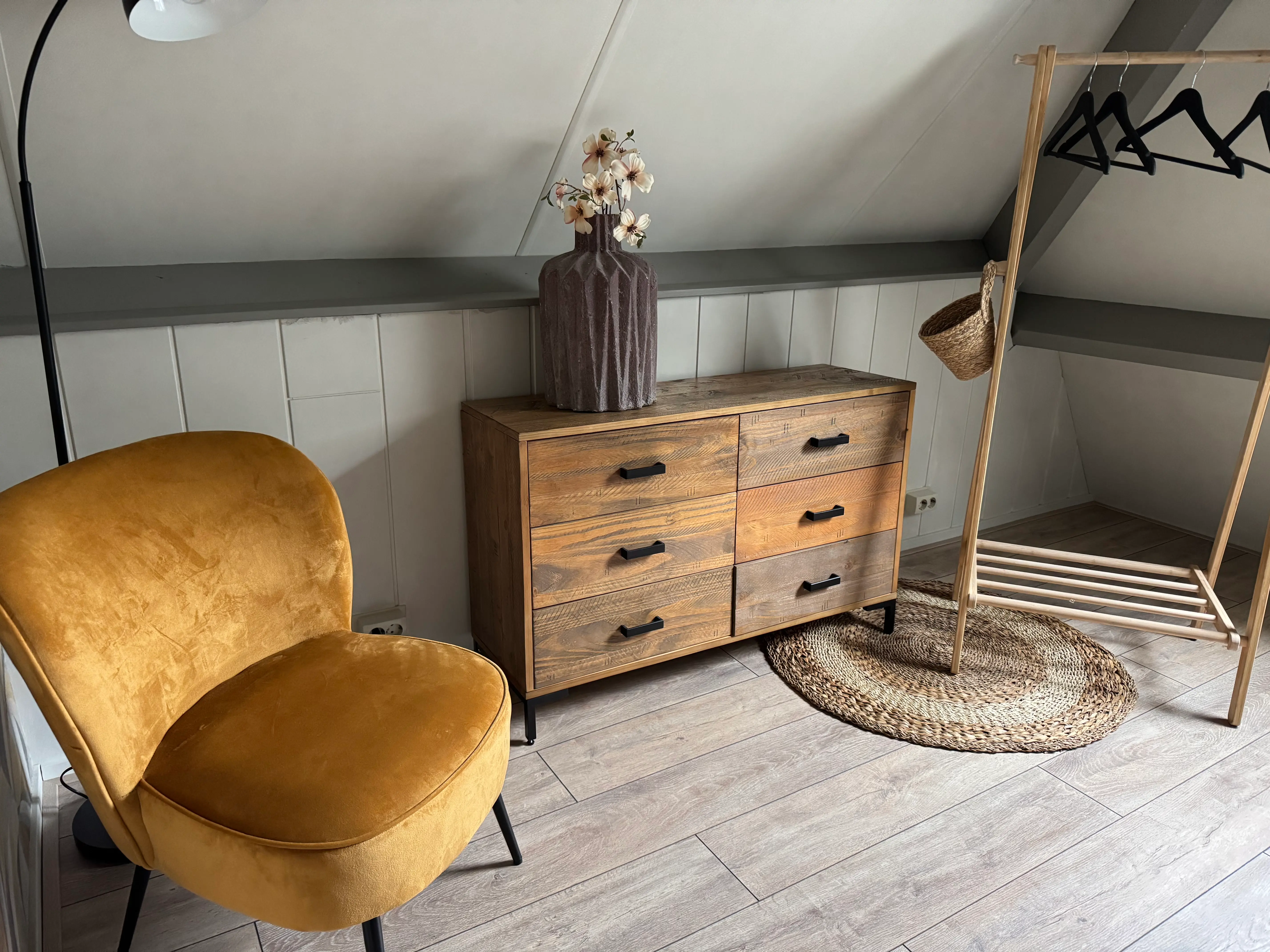 Gele fluwelen stoel naast houten ladekast en kledingrek.