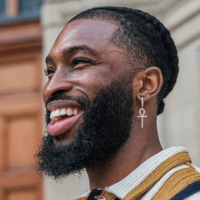 A man with a beard smiles brightly while wearing an ankh earring. He has braided hair and is set against a blurred urban background, conveying happiness.