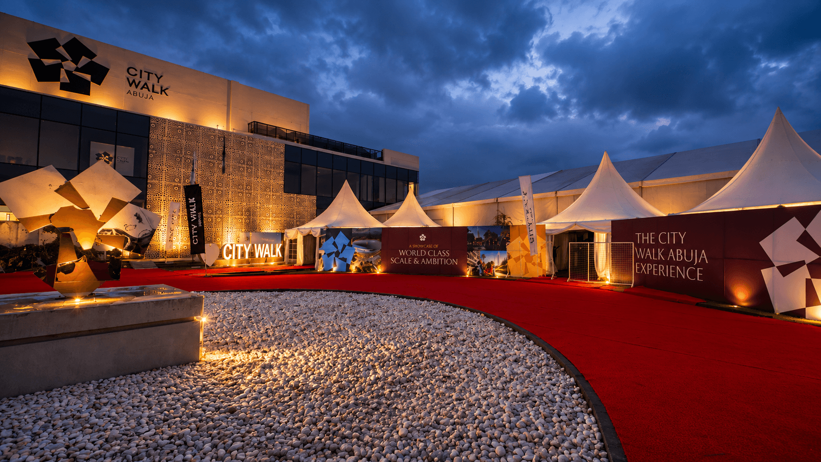 A nighttime view of the City Walk Abuja 2026 event, showcasing elegant event branding with illuminated signage, a striking modern building, decorative sculptures, and white tents set against a dramatic cloudy sky.