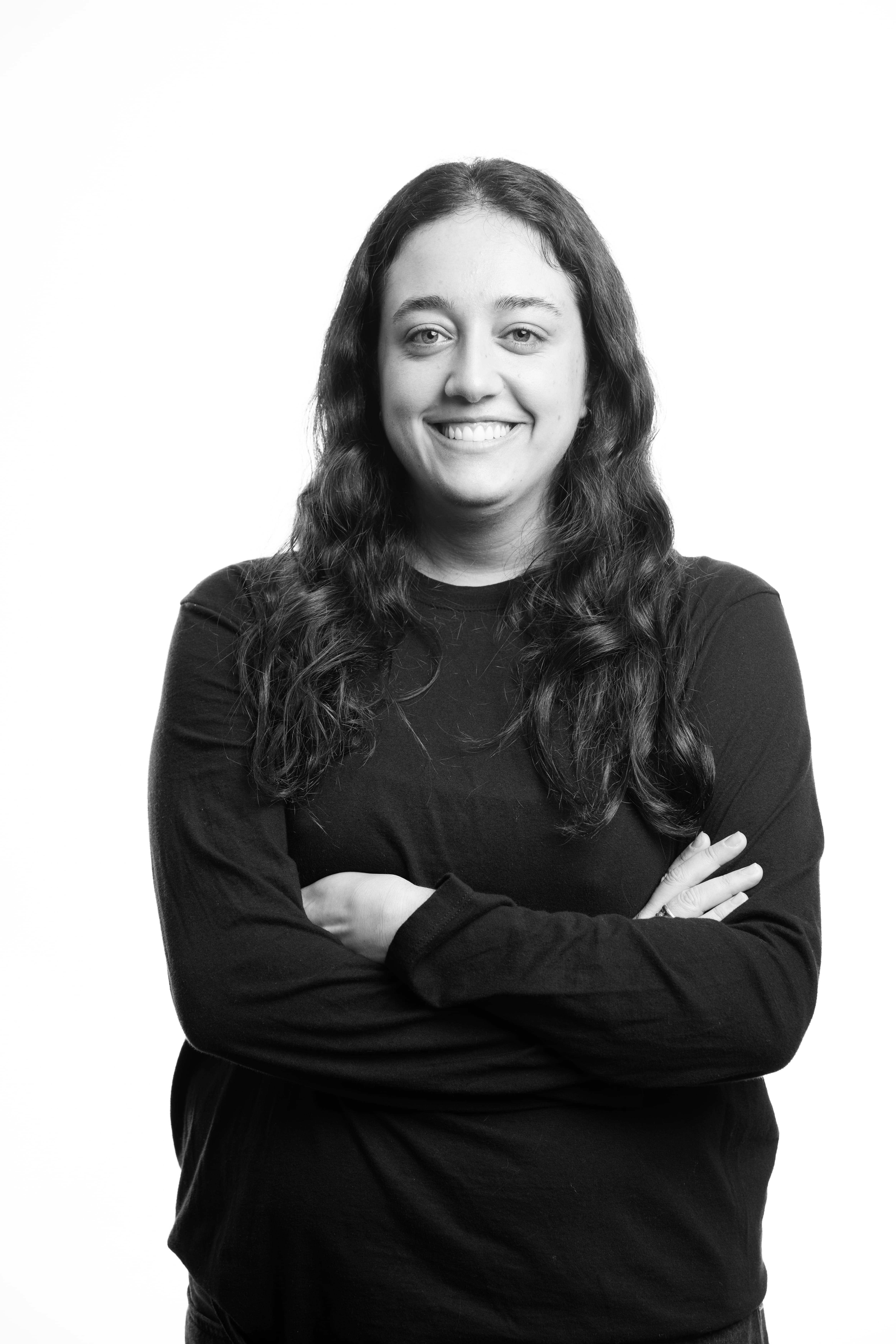 Black and white portrait of a person smiling with arms crossed, wearing a dark long-sleeve shirt. The background is plain white, conveying a confident, friendly tone.