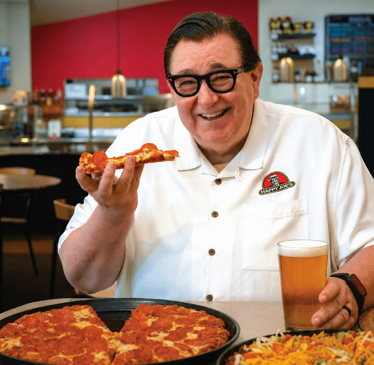 Tom Sacco holding a slice of pepperoni pizza inside a Happy Joe’s restaurant.