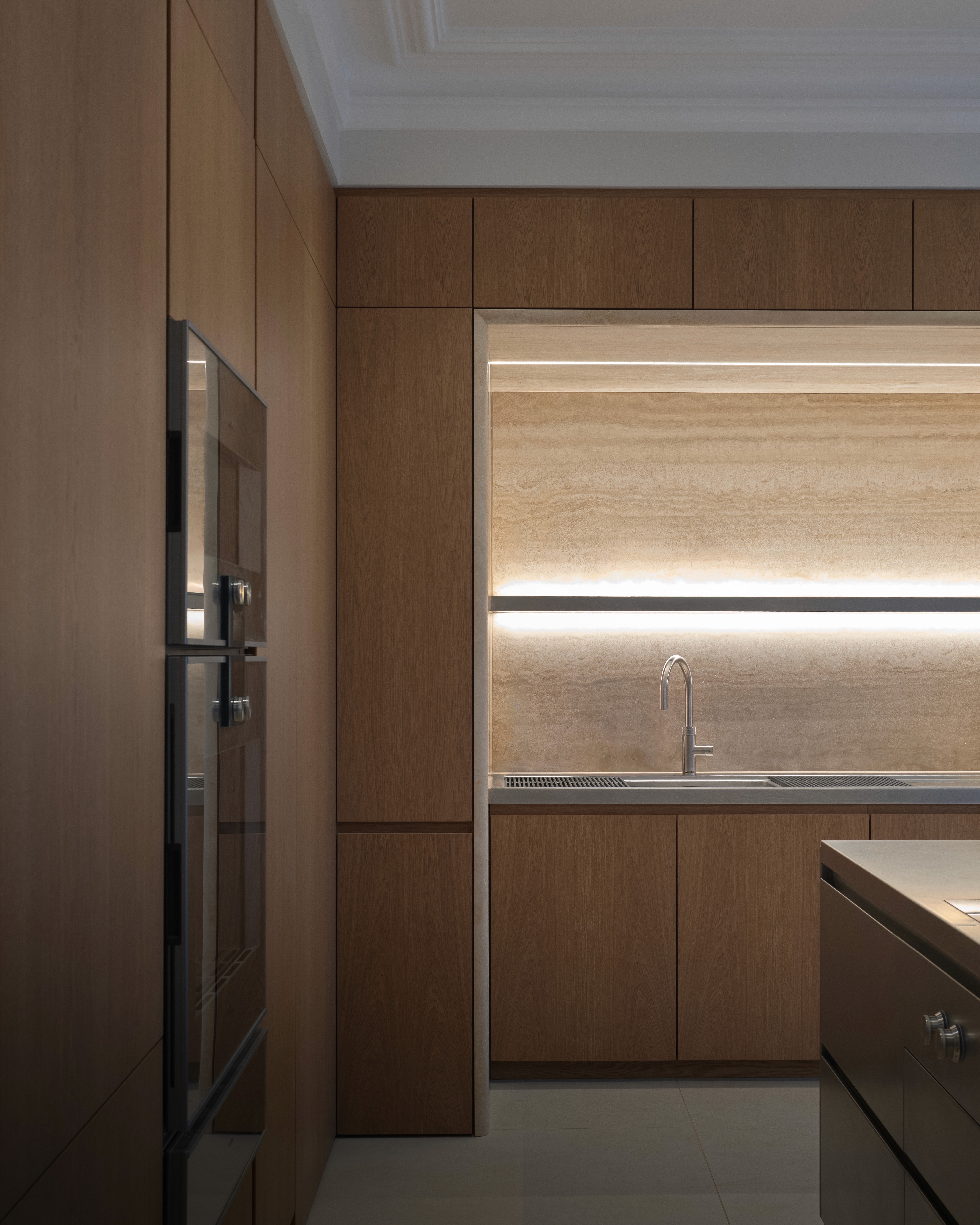 Contemporary oak kitchen cabinetry with integrated lighting and stainless steel worktop and travertine wall cladding