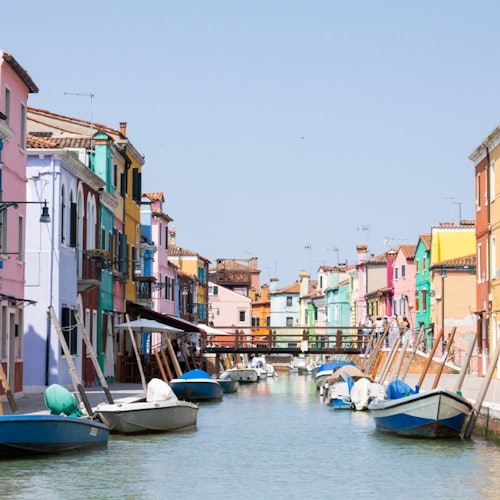 Colorful buildings line a canal with boats docked alongside on a clear day. A small bridge spans the canal in the background.