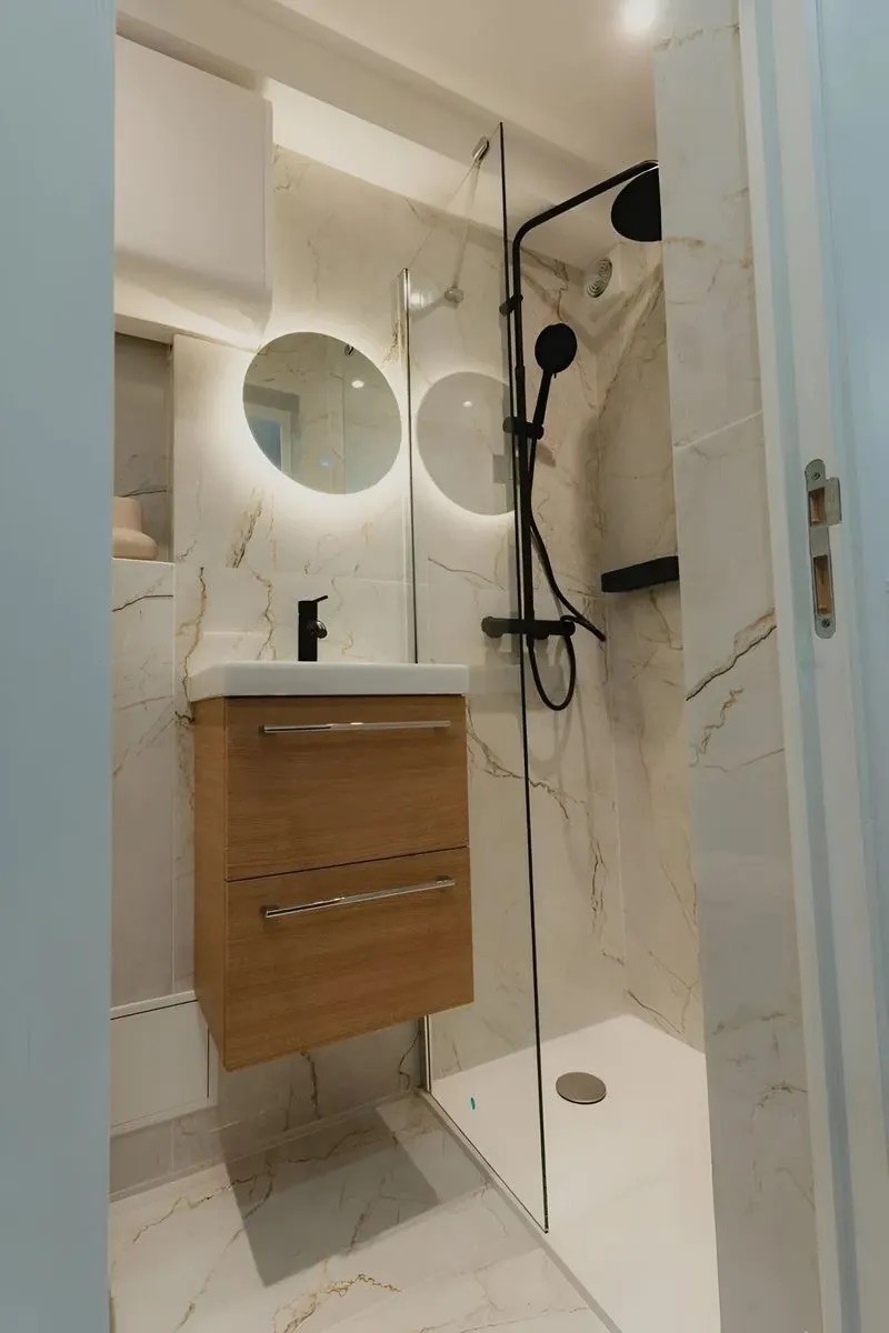 Petite salle d'eau moderne avec meuble vasque en bois clair, miroir rond rétro-éclairé et carrelage mural beige.