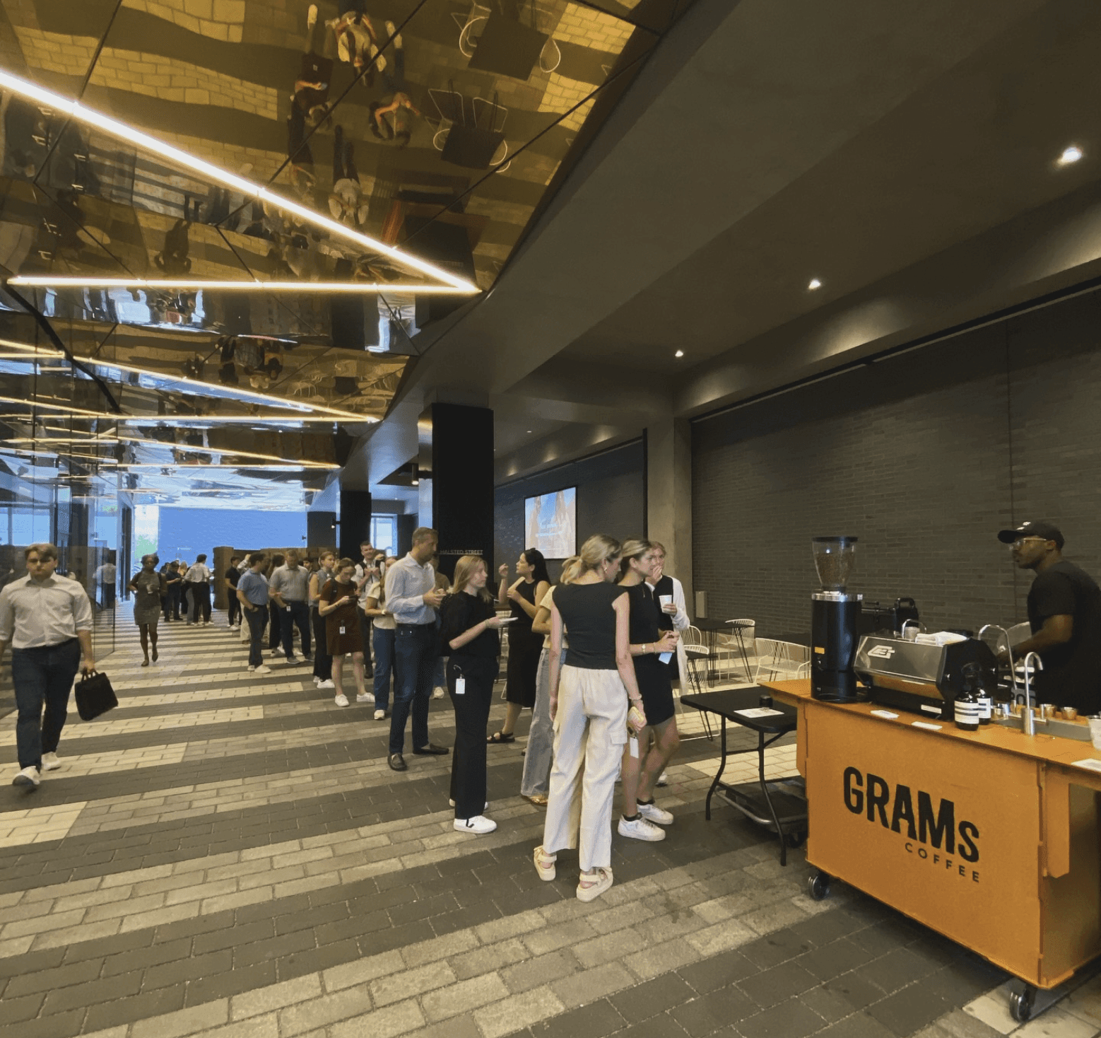 A barista at a Grams Coffee cart serves drinks to a long line of people in a modern building lobby with mirrored ceilings.