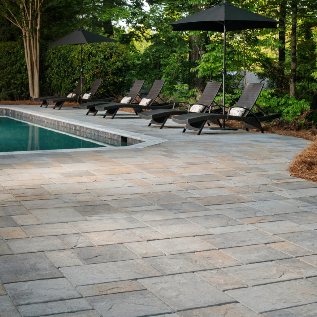 Gray and tan paver pool deck with lounge chairs and dark umbrellas beside the water.