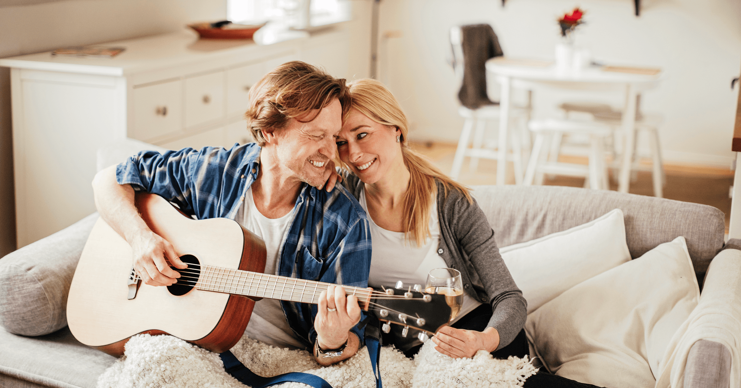 En mand spiller guitar for en kvinde på en sofa, begge smiler og nyder musikken og hinandens selskab.