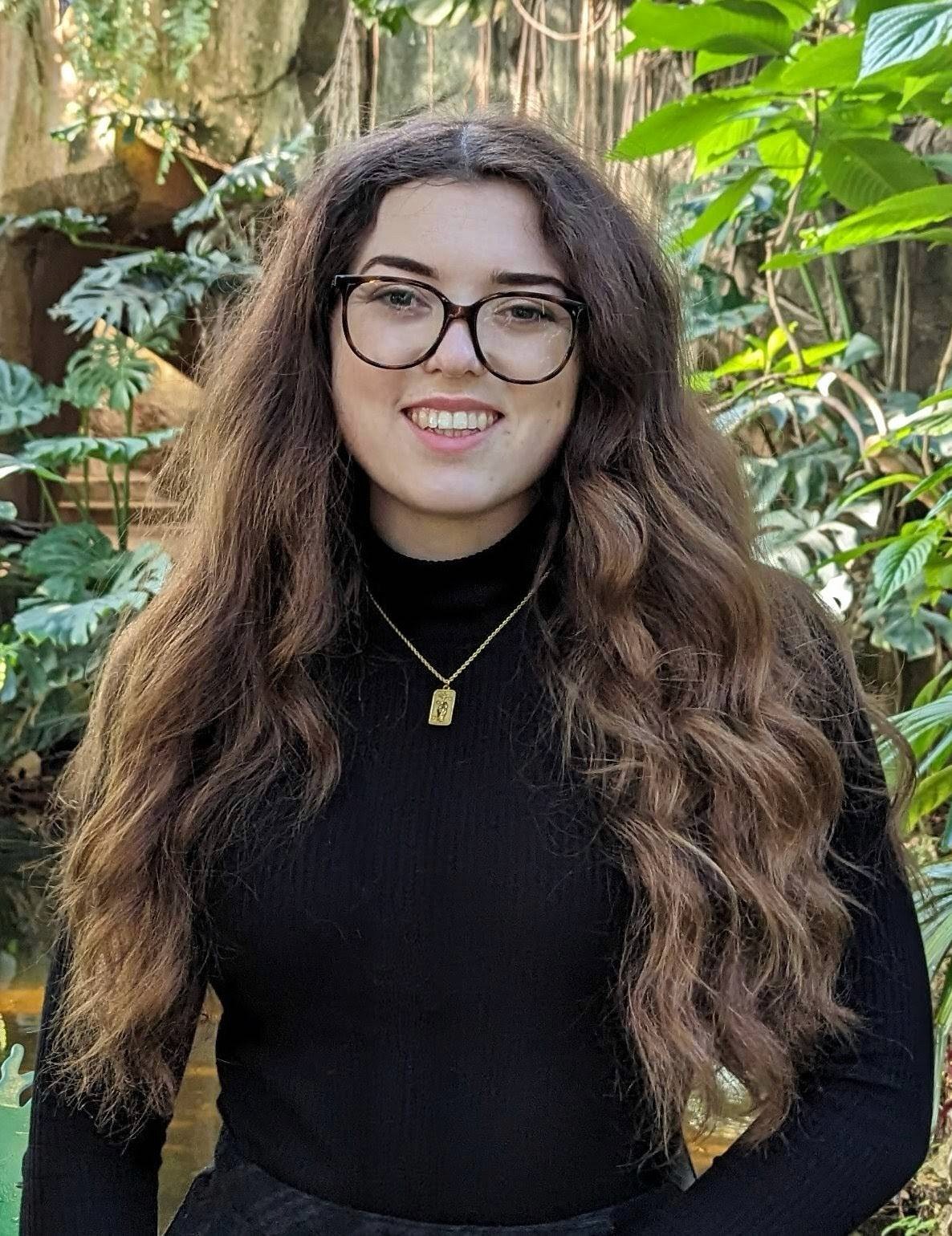 A person with curly hair and glasses smiles at the camera, seated in a cozy workspace with warm lighting.