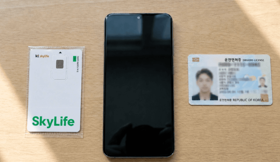 나무 책상 위에 놓인 스카이라이프 알뜰폰 유심(SkyLife 3 in 1 USIM) 카드, 검은색 스마트폰 공단말기, 그리고 대한민국 운전면허증 신분증을 나란히 배치해 둔 개통 준비 사진입니다.